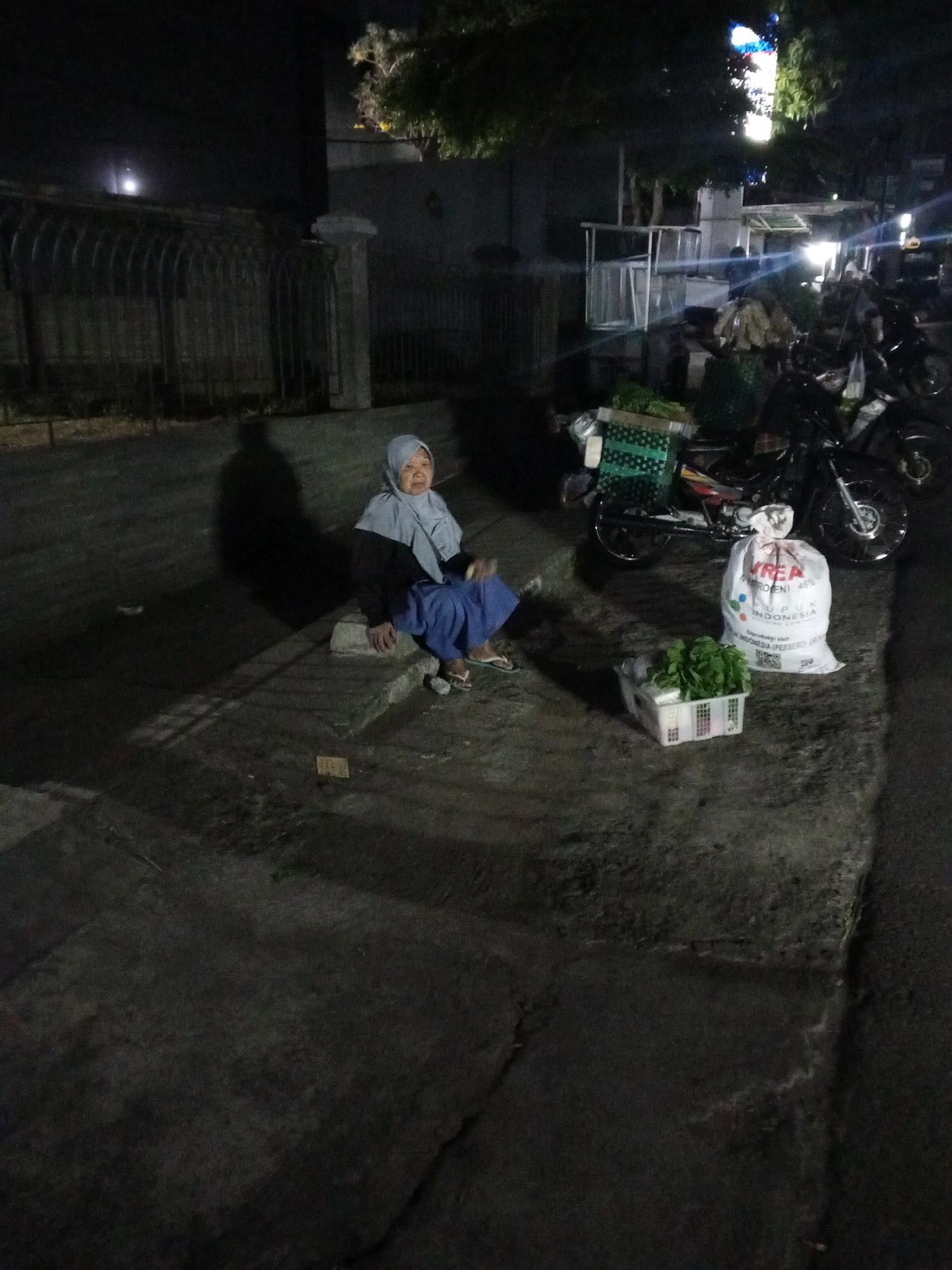 The image depicts an outdoor scene at night, likely in Gentan, Indonesia, with an elderly woman sitting on a concrete ledge. She is wearing a light blue hijab and a dark top with blue pants. To her right, a white sack with red lettering that says "KREA" and "INDONESIA" is visible, along with a basket of green leafy vegetables. Behind her, a motorcycle is parked, and there are other market stalls or structures in the background, some illuminated by lights. The ground appears to be asphalt or concrete, and a shadow of the woman is cast on it. The overall atmosphere is that of a night market or roadside stall.