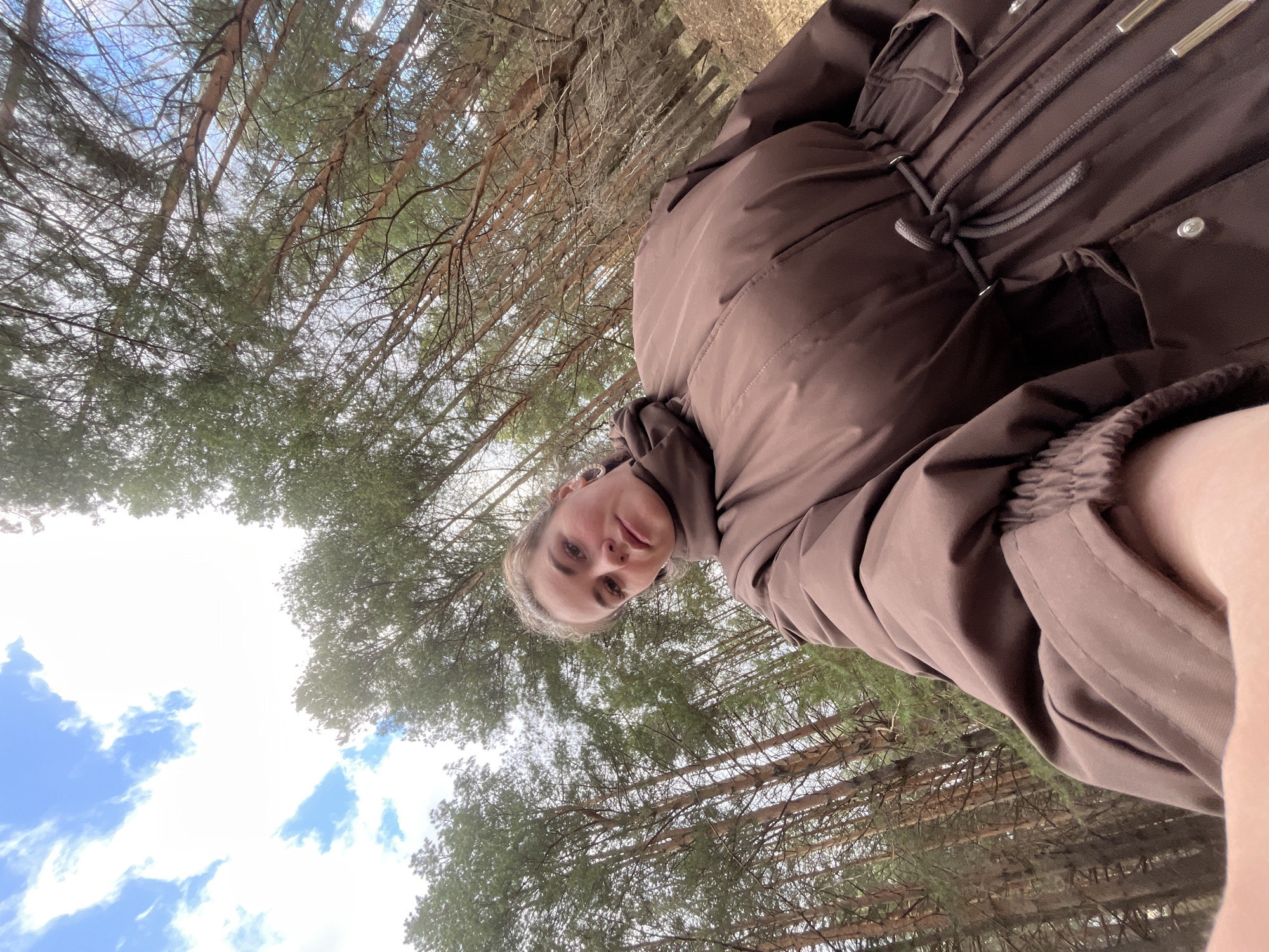 The image is a low-angle selfie of a woman wearing a brown jacket. The background consists of tall trees with green foliage and bare branches, and a sky with scattered clouds. The location is possibly a wooded area in Chaikovsky, Russia, given the context.