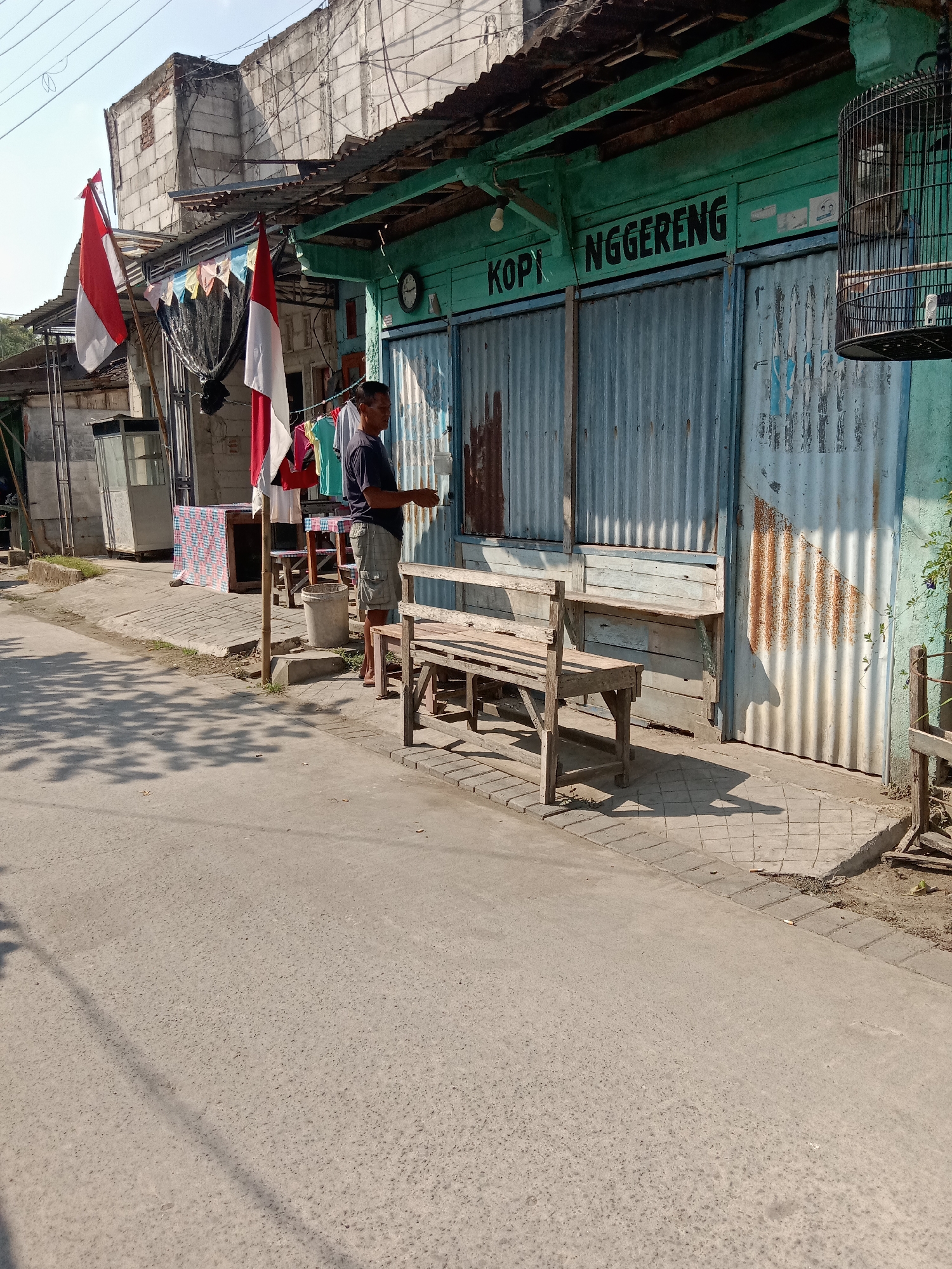The image depicts a street scene in Sugihwaras, Indonesia. A man, wearing a dark t-shirt and checkered shorts, stands in front of a shop with a turquoise facade. The shop has corrugated metal doors and a sign that reads "KOPI NGGERENG." Two Indonesian flags are displayed on poles in front of the shop, with laundry hanging on a clothesline nearby. A wooden bench sits on a paved area in front of the shop. The street is paved with asphalt, and shadows from trees are cast across it. The background shows a building with exposed brickwork and a metal roof. A birdcage hangs from the side of the shop. The environment appears to be urban, with a clear sky overhead.