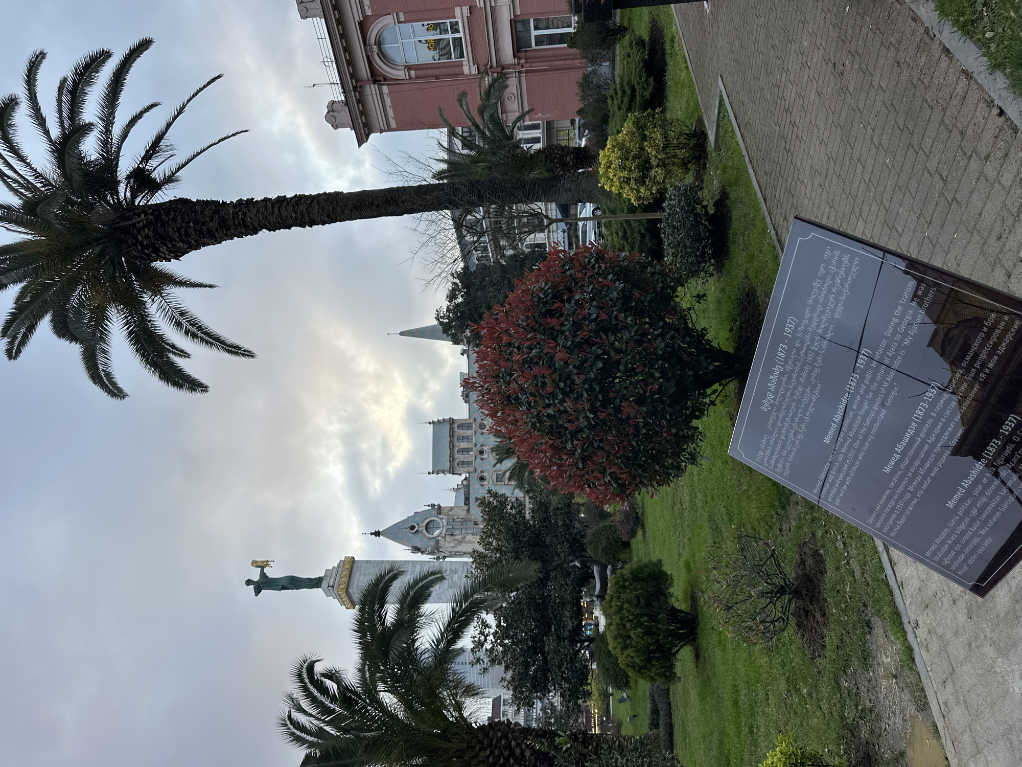Captured from a low angle in Batumi, Georgia, the photograph reveals a cityscape interwoven with lush greenery. A prominent palm tree extends from the lower left, its fronds reaching towards the sky, which is filled with gray clouds. A bronze statue of a figure stands atop a column in the mid-ground. Behind the statue and through the trees, the turquoise-tiled roofs and spires of what appears to be a historical building are visible. A red-brick building is partially visible at the upper right. In the foreground to the right is a sign providing information about Memed Abashidze. The scene is taken on a paved sidewalk next to a grassy park.