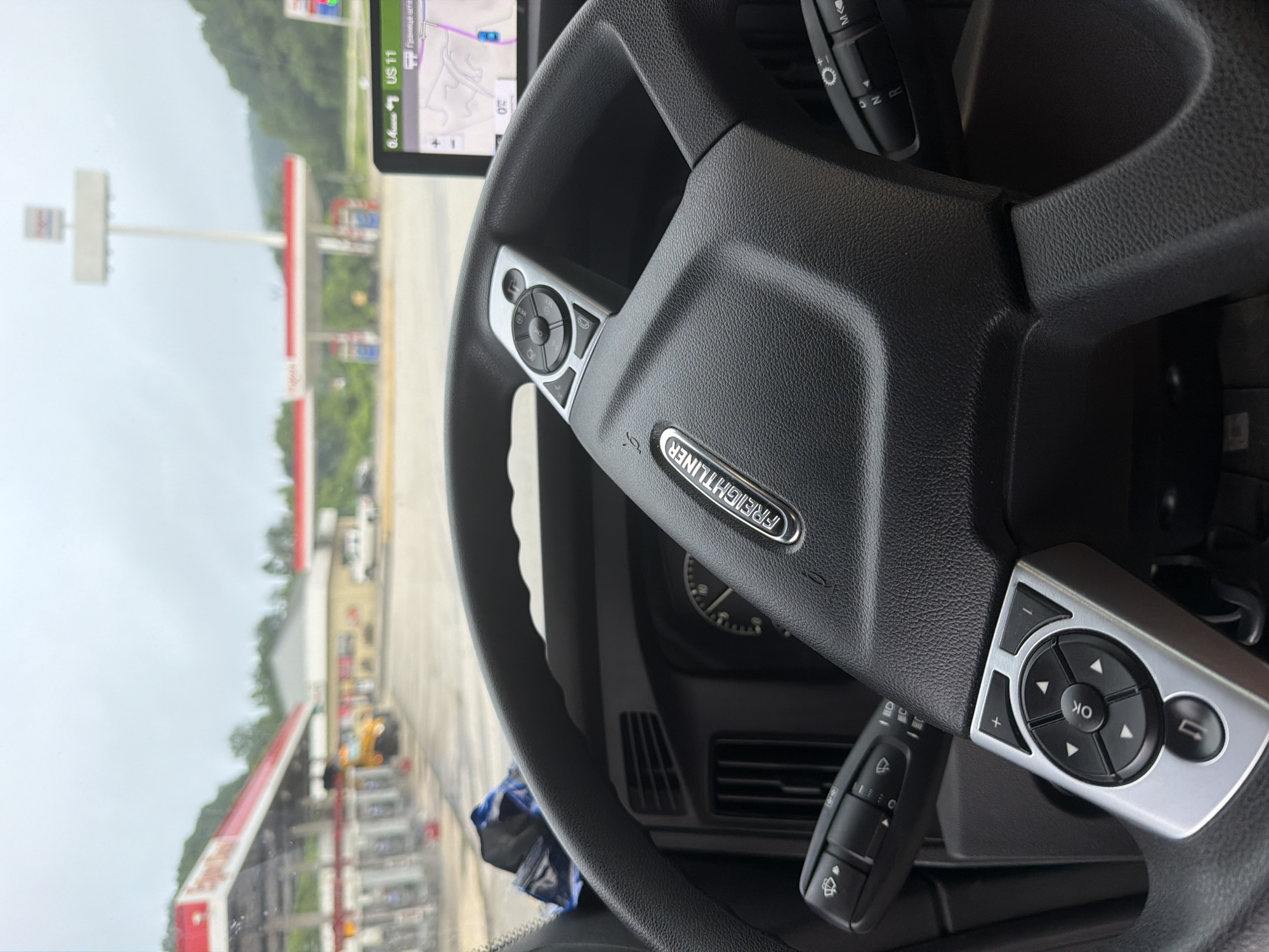 An interior shot from the driver's perspective of a Freightliner truck shows the steering wheel and dashboard. The steering wheel has various controls, including a directional pad and buttons. A GPS device is mounted on the dashboard to the right, displaying a map and traffic information. Outside the truck, a gas station with red and white awnings is visible, along with pumps and a tall pylon sign. Trees and hills form the backdrop under a cloudy sky. The location is identified as Wildwood, United States.