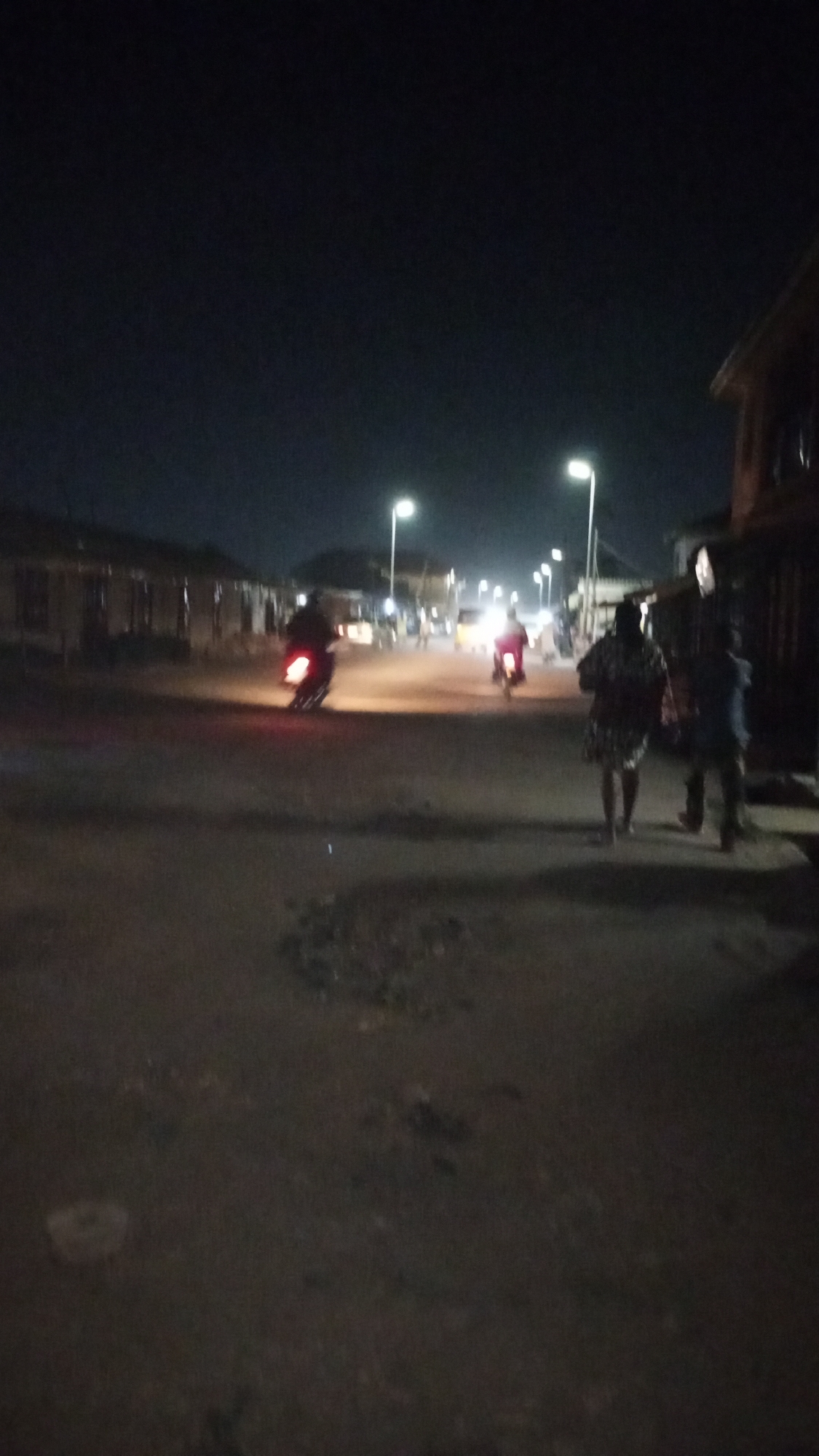 The image depicts a street scene in Sabon Tasha, Nigeria, at night. Two motorcyclists are riding down the street, illuminated by streetlights. Two people are walking on the right side of the street. Buildings line both sides of the street. The ground appears to be unpaved.