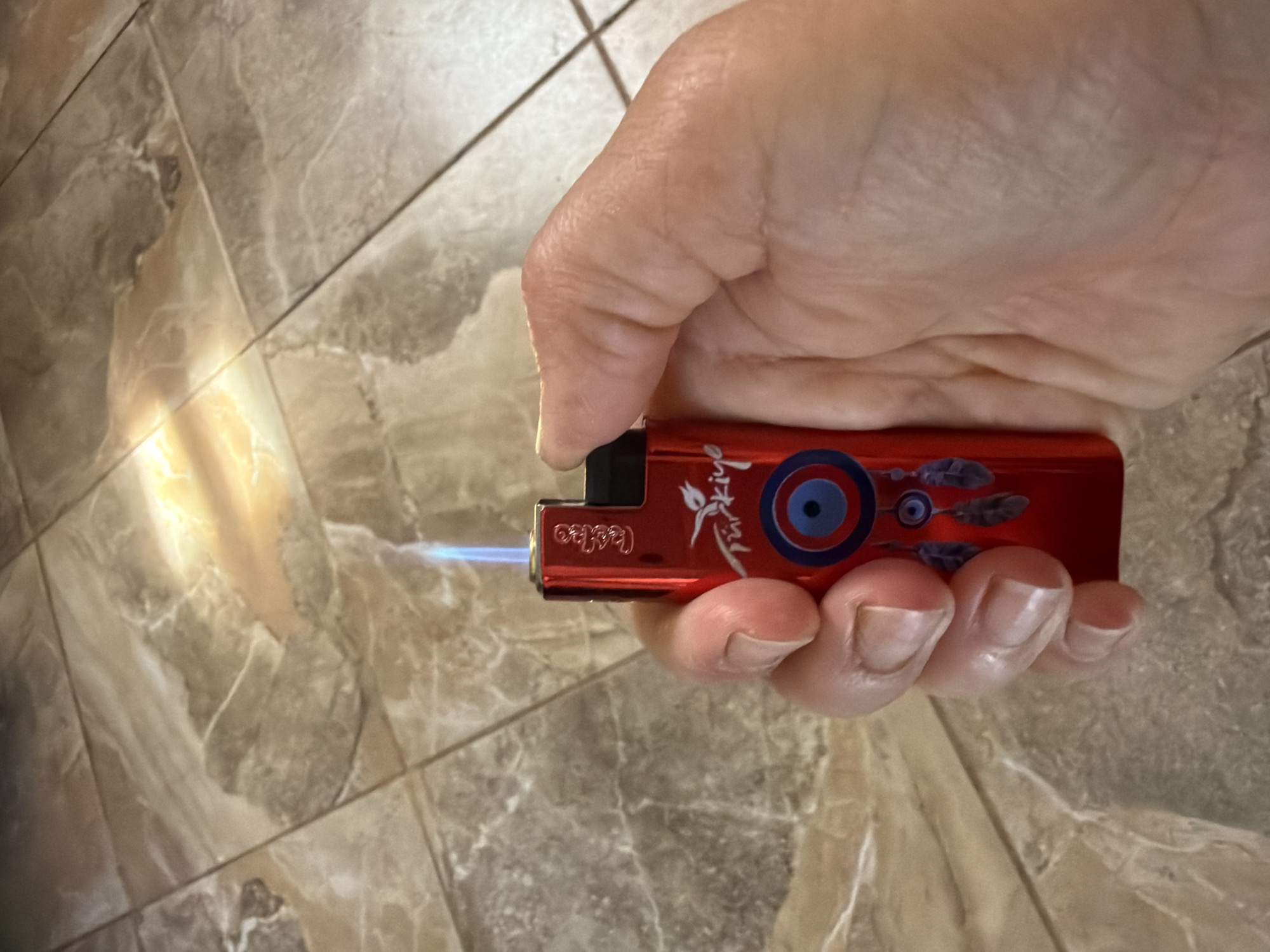 A close-up, eye-level shot shows a person's hand holding a red lighter with a blue flame. The lighter features a design with feathers and an evil eye symbol, along with the word "Türkiye" printed on it. The hand, with visible fingernails, grips the lighter, obscuring part of its body. The background consists of tiled flooring with a marble-like pattern and a bright, diffused light source illuminating the scene from the upper right. The image does not provide any information about the city of Shumki, Russia, or its location.
