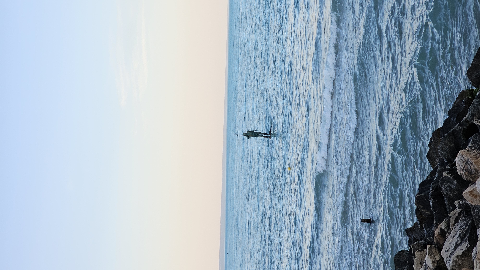 The image shows a statue of Neptune standing in the sea, near the shore. The statue is a dark color and holds a trident. The sea is choppy, with white waves breaking near the shore. The sky is a pale blue color and is more yellow near the horizon. On the right side of the image, there are some rocks. It is located in Varna, Bulgaria.