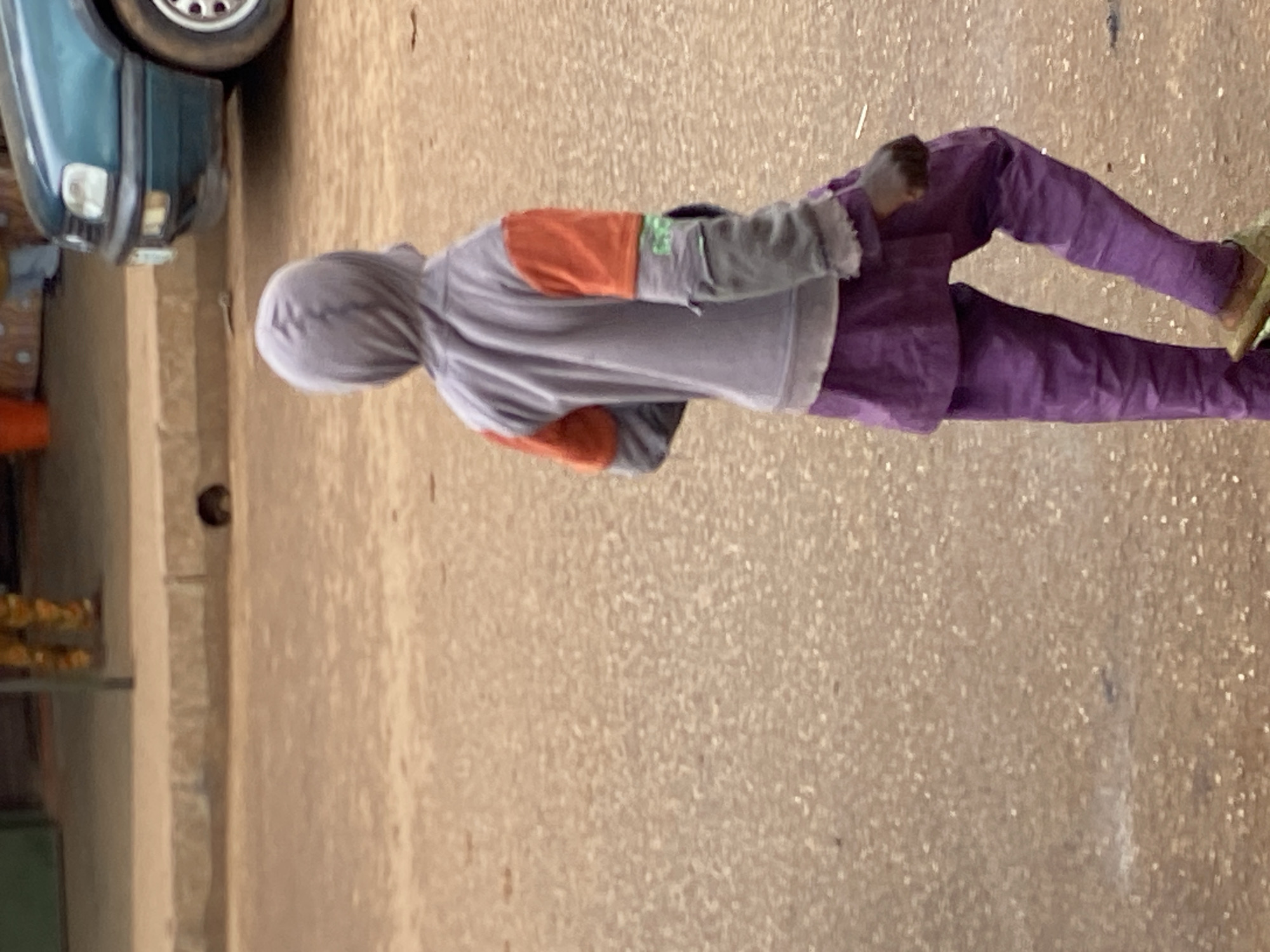 In Bukuru, Nigeria, a person wearing a grey hooded sweatshirt with orange sleeves and purple pants walks on a dirt road. A teal Volkswagen Golf is parked in the background.