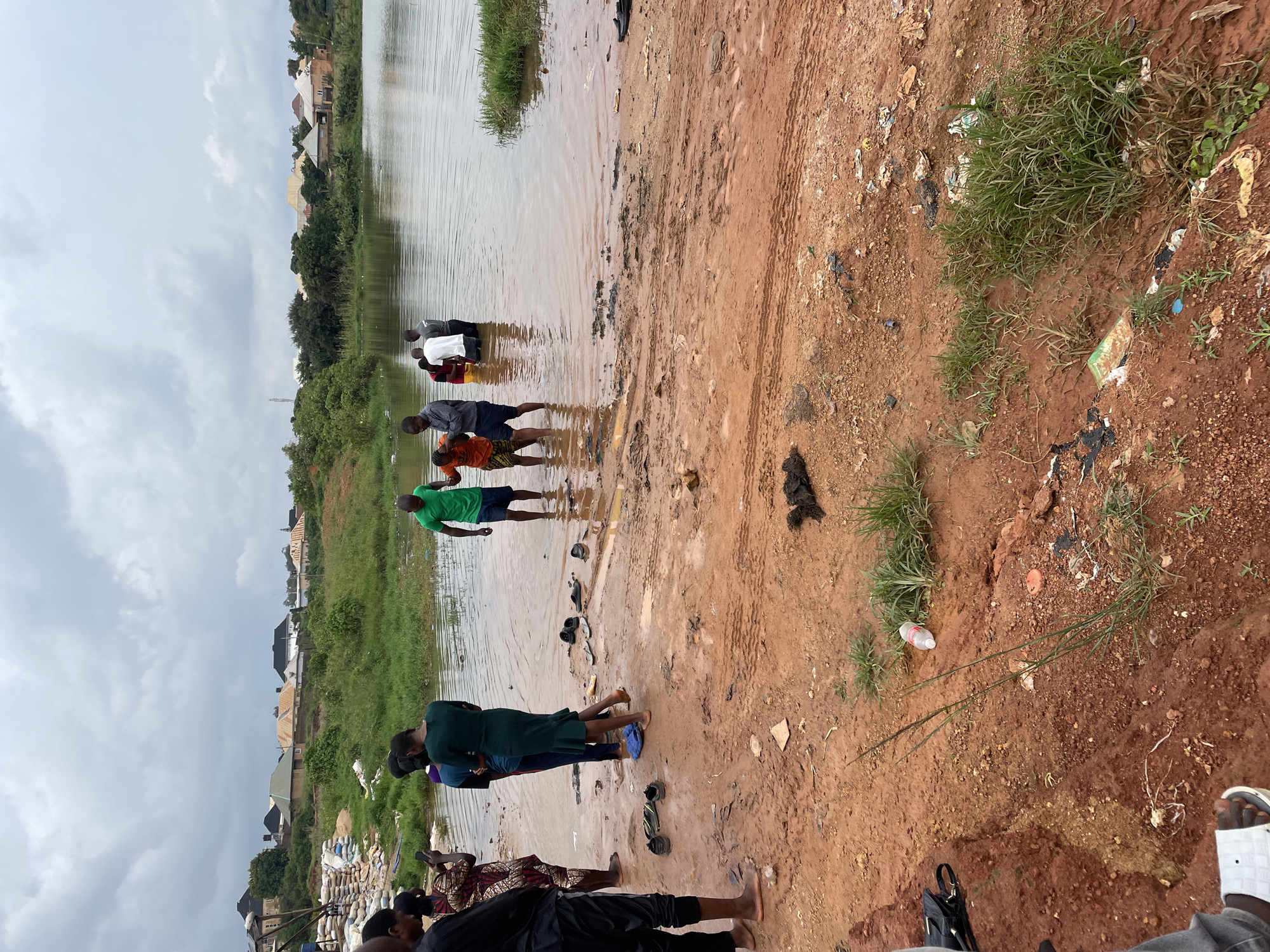 The image depicts an outdoor scene with a body of water in the background, likely a pond or lake, bordered by green vegetation and houses. In the foreground, there is a muddy, reddish-brown area with some scattered debris, including plastic bottles and trash. Several people are visible, some standing in the water and others on the muddy bank.
Specifically, the image shows a group of individuals near the water's edge. One person, wearing a green shirt, is standing in the water. To the left of this person, a woman in a green dress is also in the water, facing away from the camera. Further left, another woman in a patterned dress is standing on the muddy bank, holding a mobile phone. A man in a black shirt and pants is standing to the far left, partially visible.
In the water, towards the center, there are two men, one in a white shirt and another in a grey shirt, both facing the water. To their left, a man in an orange shirt is being assisted by another person. There are also several pairs of footwear, including sandals and shoes, scattered on the muddy ground.
The background reveals a line of houses with various roofing materials, suggesting a residential area. The sky is overcast with clouds. The scene appears to be a natural or semi-natural environment within a populated area.
Based on the context of a Jos, Nigeria location, this scene might represent a religious gathering, a baptism, or simply people interacting with a local body of water. The presence of debris suggests that the area may not be well-maintained.