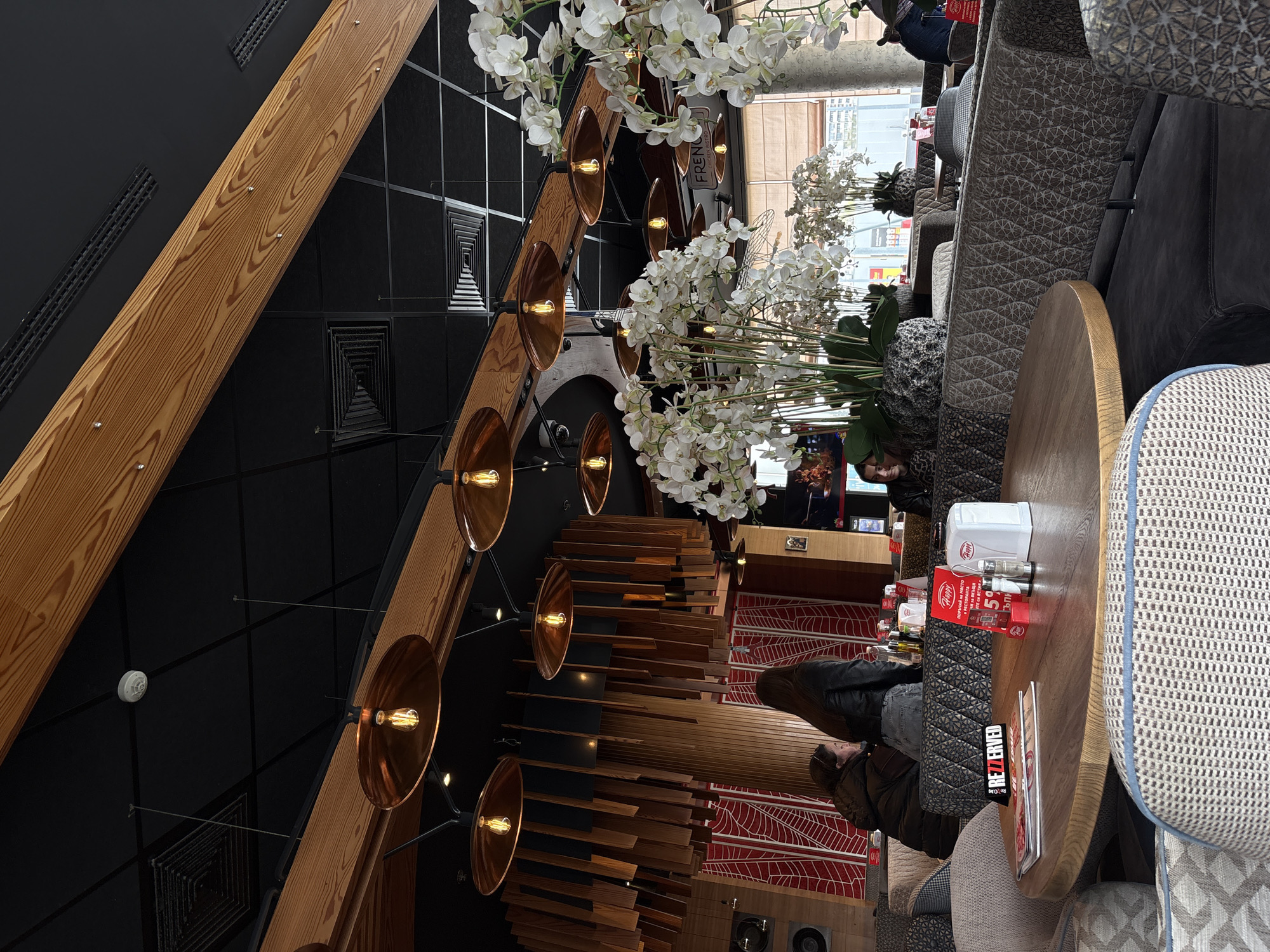 A high-angle interior shot of a restaurant in Plovdiv, Bulgaria, shows a modern, stylish space. The ceiling is a black grid with wooden beams and copper-colored pendant lights. White orchid arrangements add a touch of elegance. Down below, tables with seating areas and chairs with mixed patterned upholsteries are visible. Several patrons, including one wearing a black leather jacket, are seated and appear to be dining. Tabletop items such as napkin dispensers, salt and pepper shakers, menus, and promotional materials with Bulgarian text, including the phrase "Поръчай на място" ("Order in place") and the number "50%", are on the tables. The interior's design also includes red and white patterned carpet.