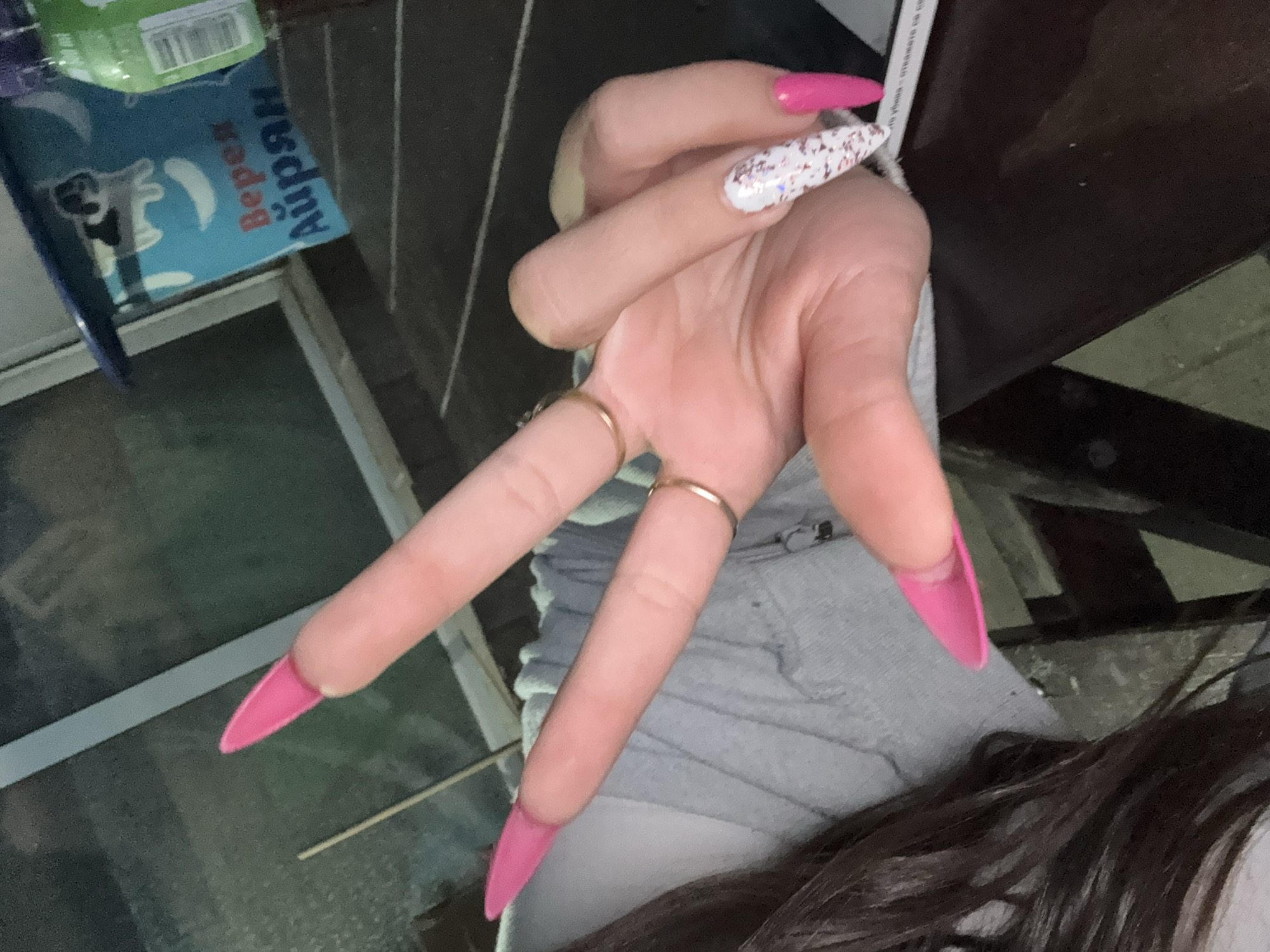 The image shows a close-up of a person's hand making a peace sign. The hand has long, bright pink stiletto-shaped acrylic nails on most fingers, with one finger sporting a white nail with glitter. Two thin gold rings are visible on the fingers. The person is wearing a light grey hooded sweatshirt. The background is blurred but appears to be an outdoor setting, possibly a patio or garden area, with a table and some containers visible. One container has text in Cyrillic script. The image composition suggests the photo was taken by the person themselves. There are no indications of the city of Malo Konare, Bulgaria in the image.