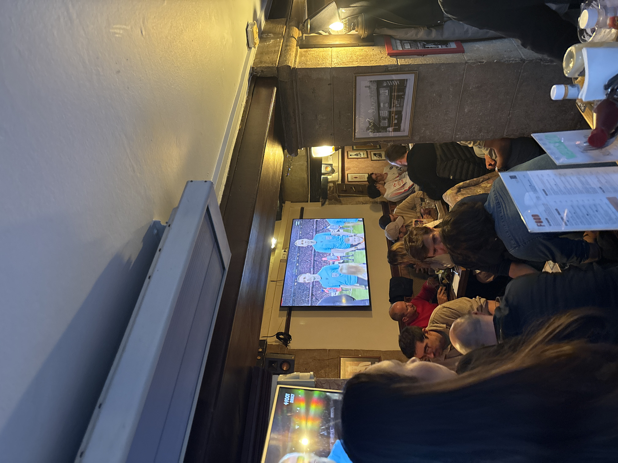 A busy interior scene, possibly a pub or sports bar, shows a group of people watching a sports match on a large television screen. The TV displays a soccer/football game with players in blue jerseys. The environment appears to be dimly lit. Various individuals are seated at tables. Some are focused on the screen, others engage in conversation or look at menus. Bottled condiments and drinks are visible on the tables. The architecture includes stone columns and framed pictures on the wall. A second television screen sits lower down. Based on the information given, the location is supposedly in Paris, France, although definitive proof is absent from the imagery.