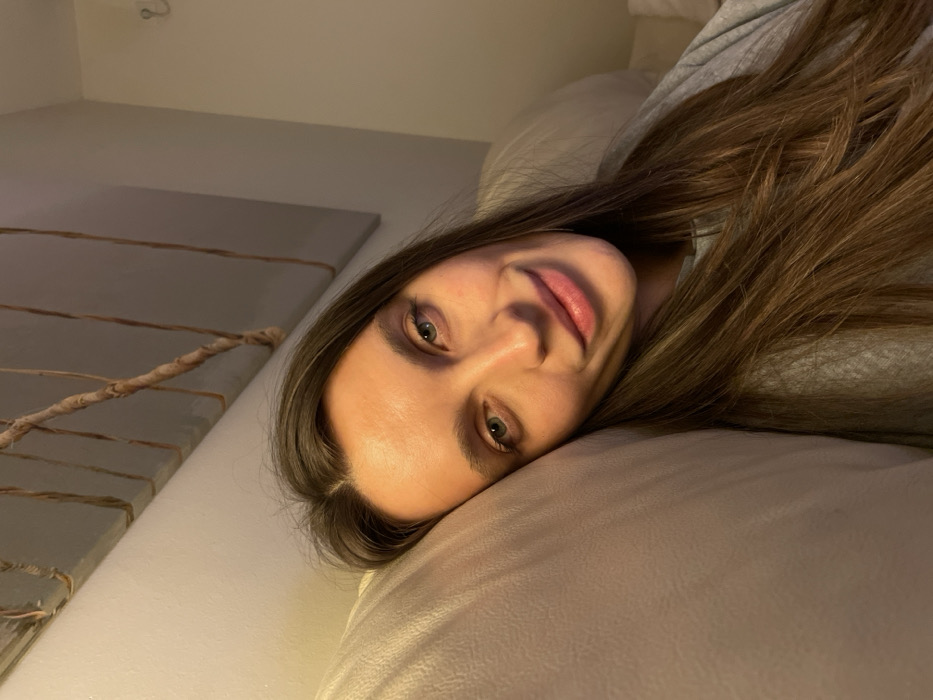 A woman with long brown hair rests her head on a white pillow. Her skin tone is fair, and she looks directly at the camera with light green eyes. The background is a dimly lit room, featuring a gray textured object adorned with intertwined rope. A wall is visible in the upper portion of the frame. The overall composition is a close-up shot, focusing primarily on the woman's face and hair.