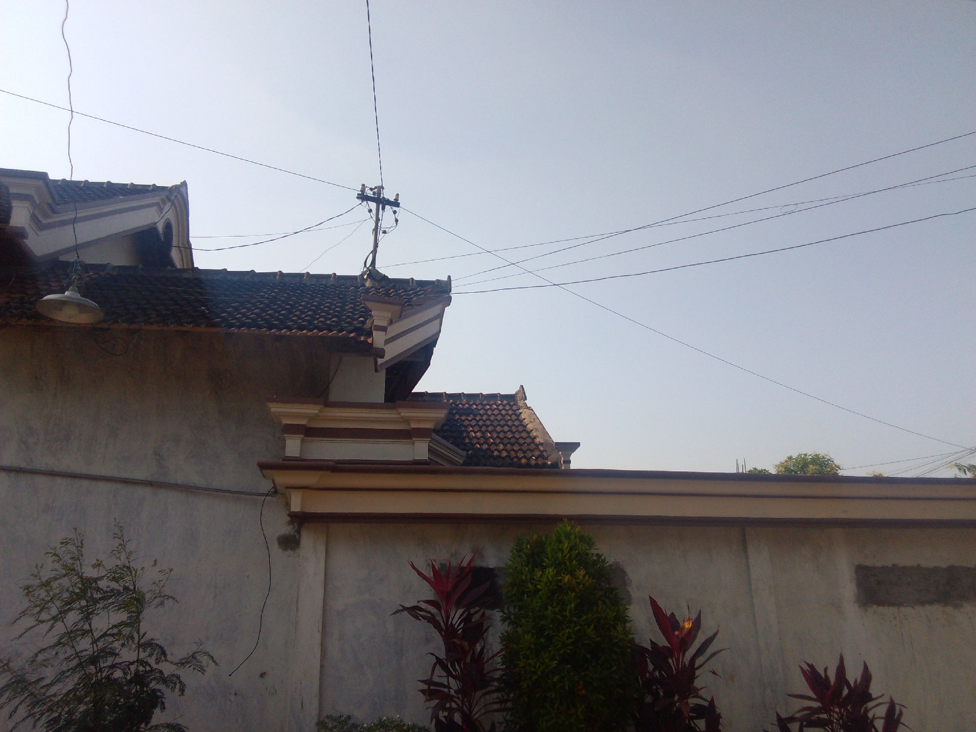 A low-angle, medium shot captures the exterior of a residential building and its surrounding environment under a clear, pale blue sky. The building features a tiled, brown roof with decorative white and brown trim along the eaves. A simple, metal pendant light fixture hangs from the roof on the left side of the frame. Electrical wires crisscross the sky, originating from a utility pole visible on the rooftop in the upper right quadrant. The building's walls are a neutral gray, with a lighter colored horizontal band running across the middle.
In the foreground, on the left, a sparse, leafy green tree with thin branches is partially visible. To the right, closer to the building's wall, are several ornamental plants with dark red, elongated leaves, and a small, dense green shrub. The overall scene suggests a residential setting in Karangan, Indonesia, characterized by simple architecture and a clear, bright sky. There are no people or discernible actions within the frame.