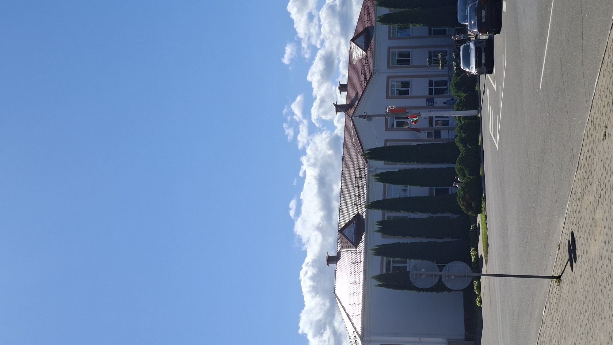The image depicts the exterior of a building in Myadzyel, Belarus, on a clear day with scattered clouds. The building is a multi-story structure with a white facade and a reddish-brown tiled roof. Several tall, slender evergreen trees line the front of the building, interspersed with manicured bushes. In front of the building, there are flags flying from poles. The foreground shows a paved road and a sidewalk, with cars parked and driving on the road. A street signpost with two circular signs is visible on the left side of the image. The overall scene suggests a civic or administrative building, possibly a school or government office, located in an urban or town setting.
