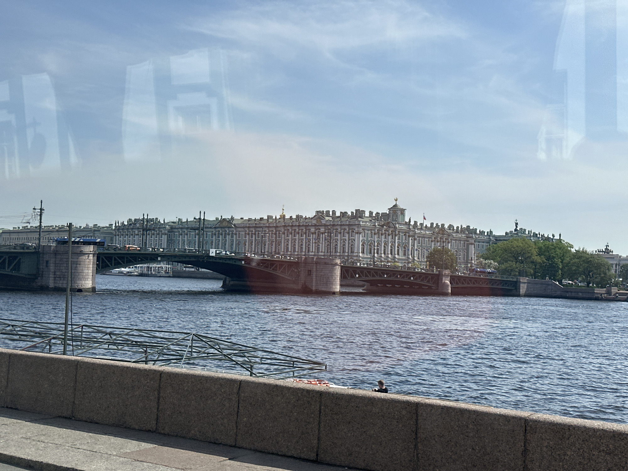 The image shows a view of the Neva River in Saint Petersburg, Russia, with the Winter Palace and the Palace Bridge visible in the background. The Neva River is the most prominent feature, exhibiting slight wave action. A gray stone embankment wall runs along the bottom of the image, in front of which is a partially visible metal structure, and a solitary figure of a person seated at the water's edge. Above the embankment, the Palace Bridge spans the river, with traffic moving across it. Beyond the bridge, the Winter Palace, characterized by its ornate architecture, occupies a significant portion of the background. The sky above is bright with a few wisps of clouds. The foreground contains reflections, possibly from a window.