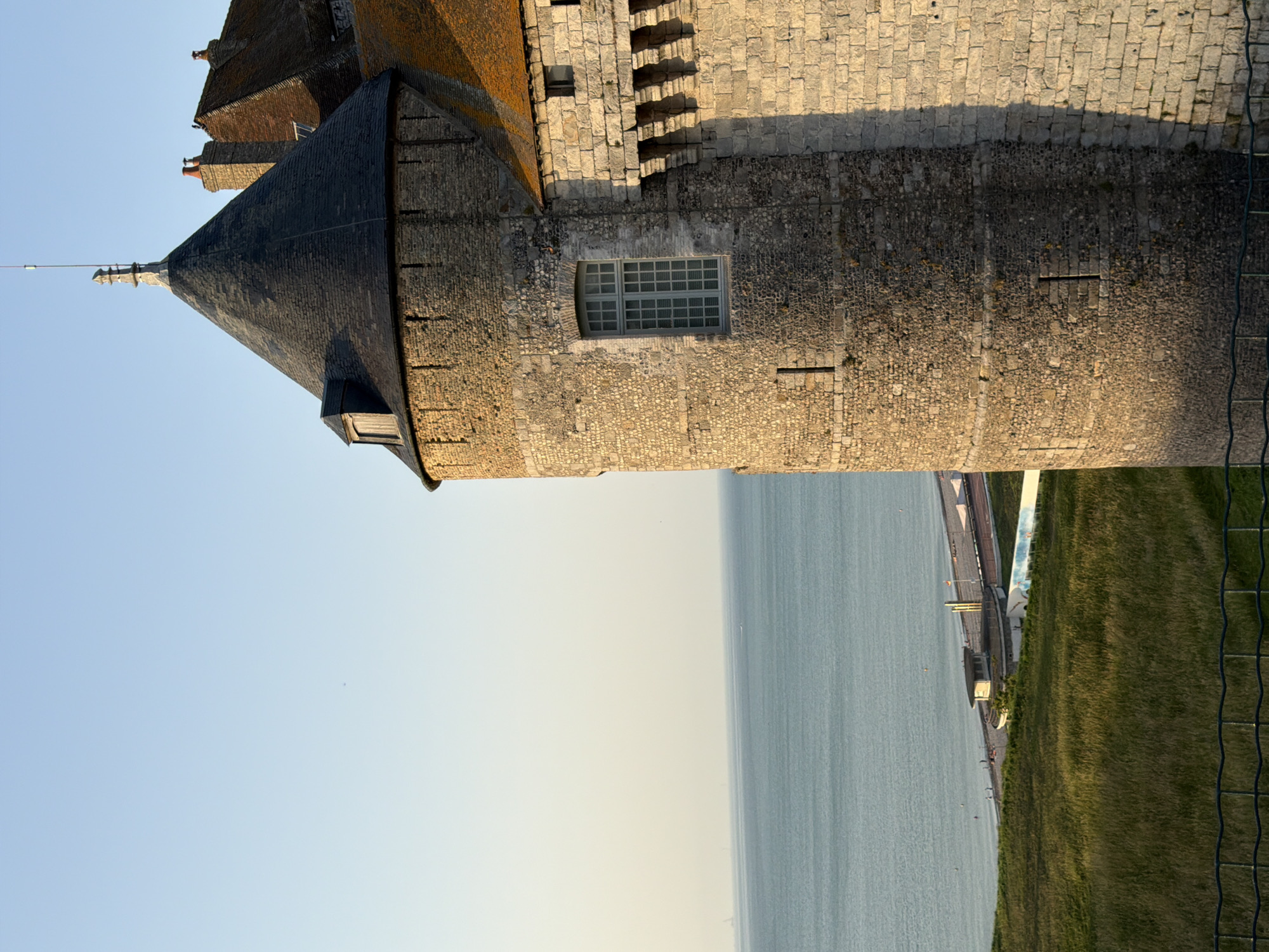 The image depicts a stone tower and adjacent building, likely part of a historic structure, with a conical slate roof on one section. The stonework of the tower is rough and uneven, composed of small, irregular stones. A multi-paned window is visible on the tower's face. The building appears to have a red tiled roof in some sections and brick chimneys. The scene overlooks a calm body of water, possibly the sea, with a clear, pale blue sky above. Along the water's edge, a promenade is visible with some sparse greenery and what appears to be a small building or structure. Further to the right, a grassy slope leads down towards the water, with a dark metal fence in the foreground. The lighting suggests it is either early morning or late afternoon, casting soft shadows. The overall atmosphere is tranquil. No people are clearly discernible as subjects, though there are distant figures along the promenade. The location is identified as Dieppe, France.