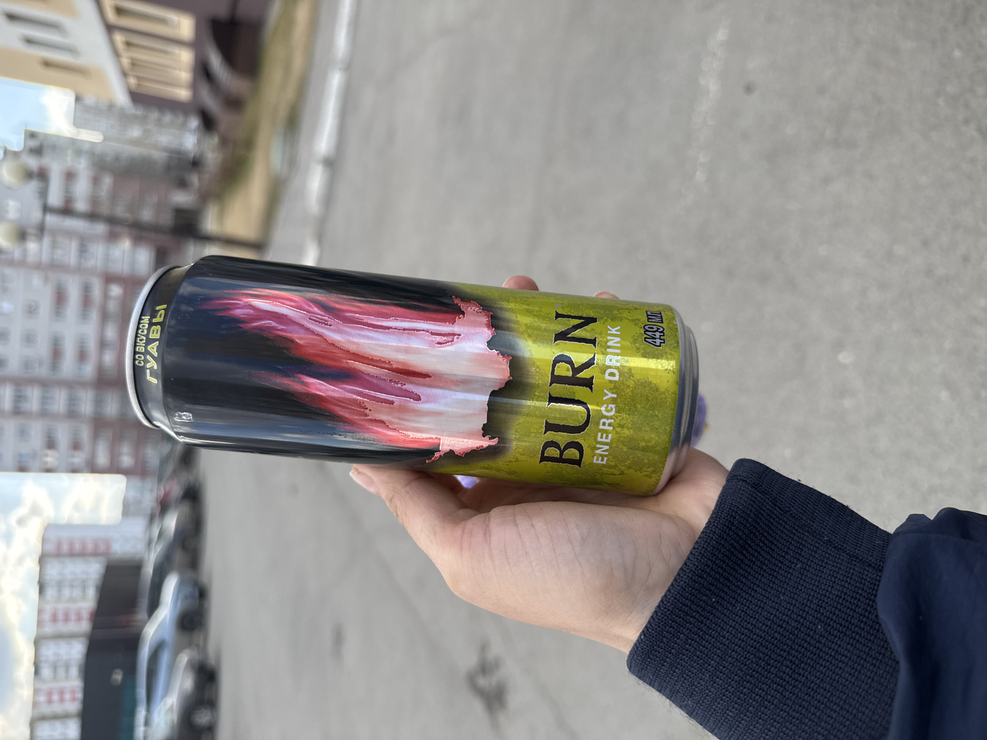 A close-up, rotated shot captures a hand holding a "BURN" energy drink can outdoors in what appears to be an urban setting in Tobolsk, Russia. The can features a striking design: a black background with a dynamic red and pink flame-like graphic, which transitions into a bright lime green section. The brand name "BURN" is prominently displayed in large white letters on the green area, with "ENERGY DRINK" printed below it. The volume "449 МЛ" (449 ml) is also visible. Further text in Cyrillic reads "СО ВКУСОМ ГУАВЫ", translating to "WITH GUAVA FLAVOR". The hand holding the can wears a navy blue jacket with a ribbed cuff.
The background is softly blurred, showing multi-story buildings, consistent with residential or commercial architecture, lining what appears to be a street or parking area. Faint outlines of parked cars are discernible. The ground beneath is a light-colored asphalt or concrete. The scene is illuminated by natural daylight, suggesting a clear or partly cloudy day. The image captures a casual moment with the energy drink against an everyday urban backdrop.