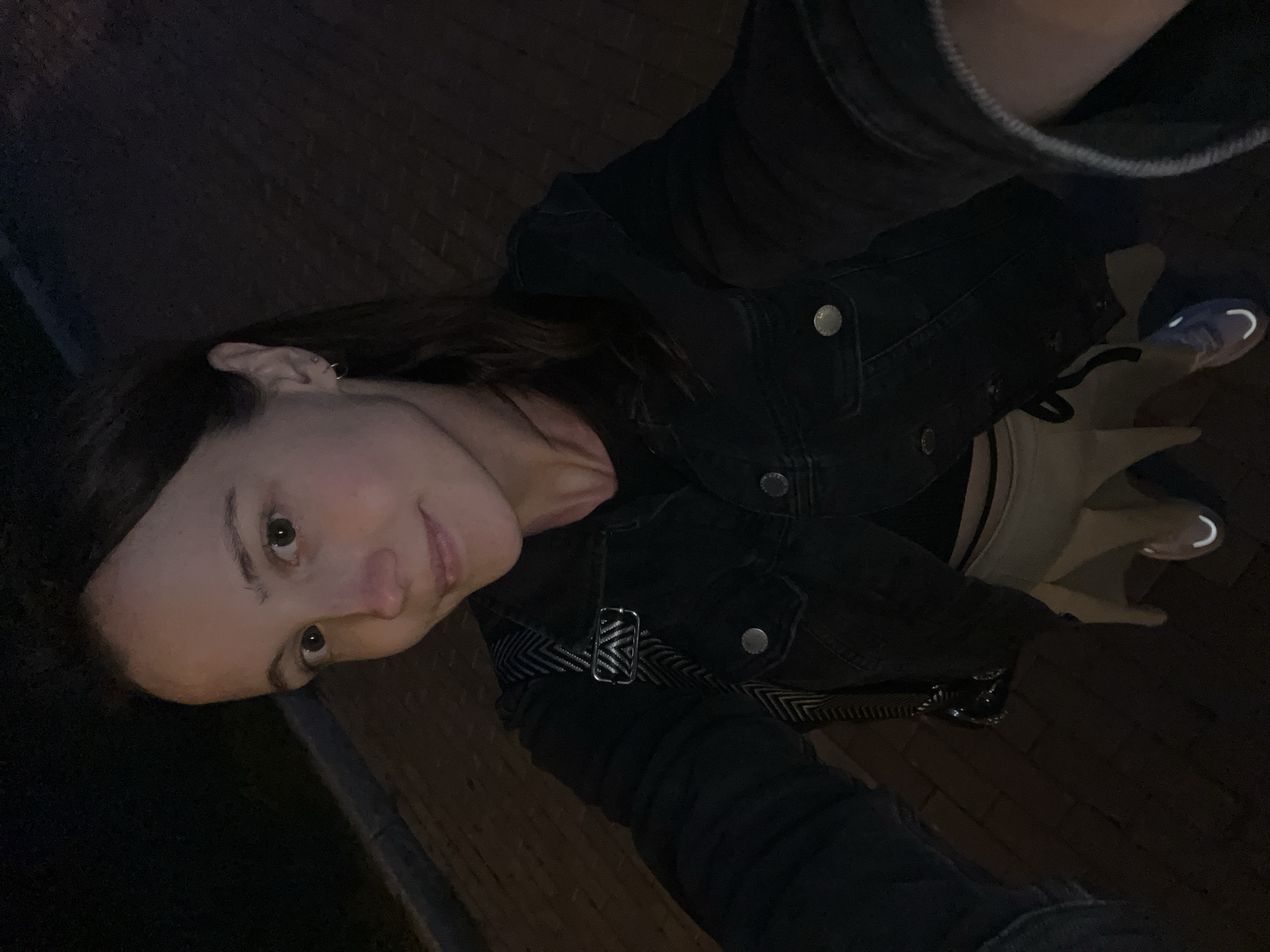 A young woman is taking a selfie outdoors at night. She has brown hair and is wearing a dark denim jacket over a black top and a tan skirt. A crossbody bag with a patterned strap is visible. She is standing on a paved surface, possibly a sidewalk or pathway. The background is dark, suggesting it is nighttime in an urban or suburban environment. The image was taken in Novokuybyshevsk, Russia.