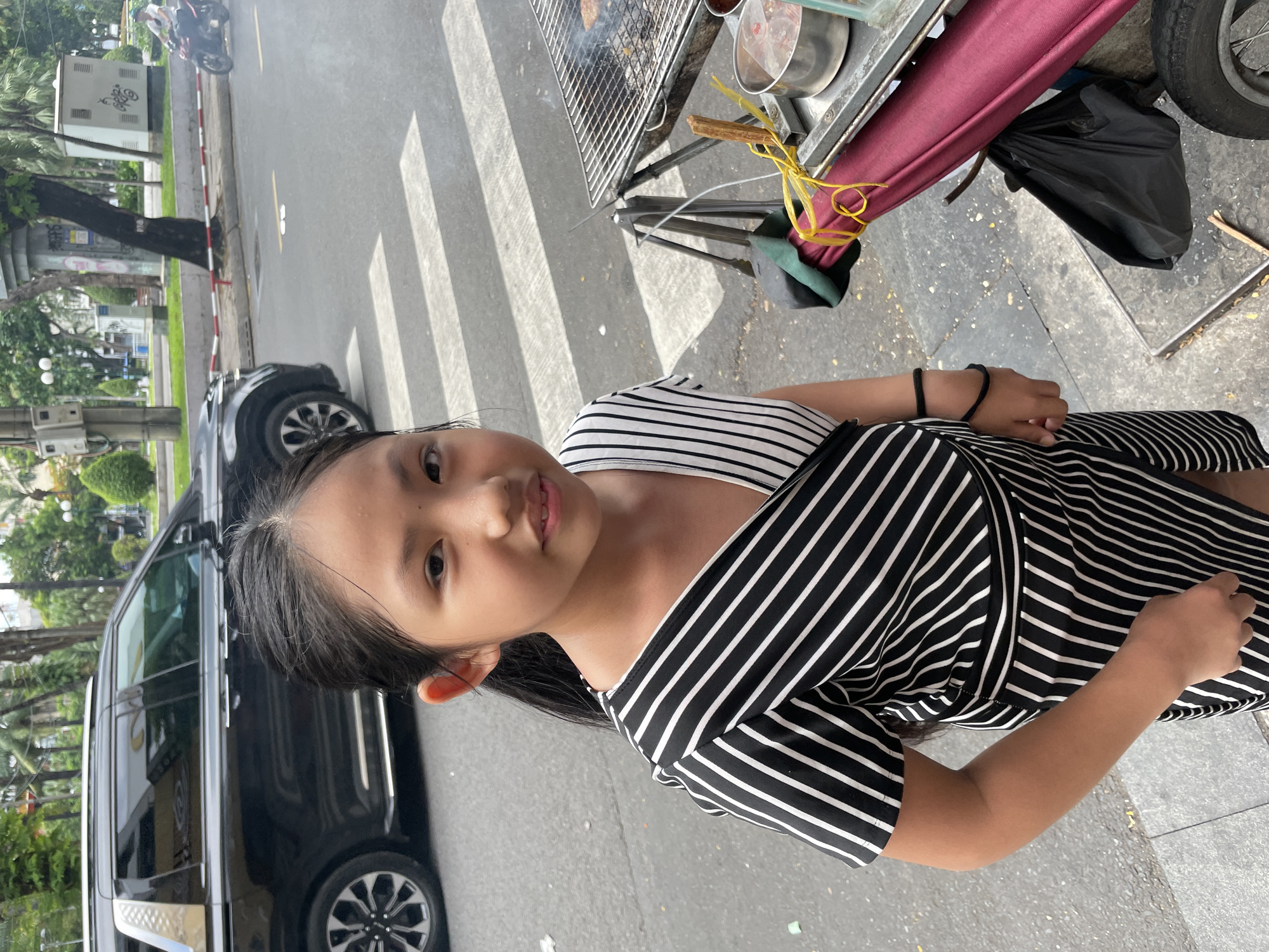 A young girl with dark hair, wearing a black and white striped dress, smiles at the camera. She is positioned to the left of the frame, with her face and upper body visible. Behind her, a black car is parked on the side of a street, with its front wheel turned slightly. The street appears to be paved, and a pedestrian crossing with white stripes is visible in the background. To the right of the girl, a food cart with a metal grill is set up. Smoke is rising from the grill, suggesting food is being cooked. The cart is covered with a red awning. A black trash bag is tied to the cart. The overall setting suggests an outdoor urban environment, possibly a street food scene in Thủ Đức, Vietnam. The lighting indicates it is daytime.