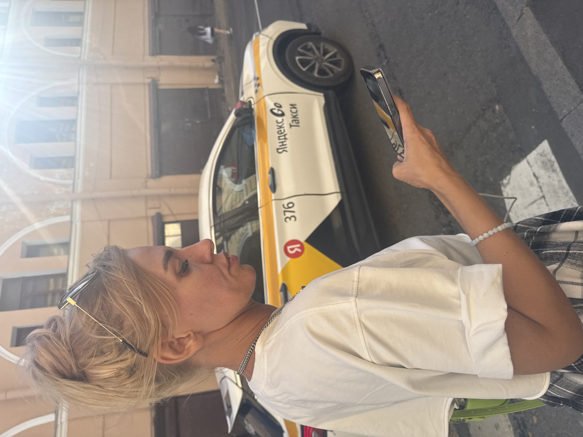 A blonde woman stands on a city street in Saint Petersburg, Russia, looking at her phone. She is wearing a white t-shirt, a plaid skirt, and a silver chain necklace. Sunglasses are perched on top of her head. In the background, a white taxi with yellow and orange markings is parked. The taxi has the Yandex Go logo on it. The woman is holding a silver smartphone in her right hand. The street is paved with asphalt, and there are buildings with arched windows in the background. The sun is shining brightly, creating lens flare in the upper right corner of the image.