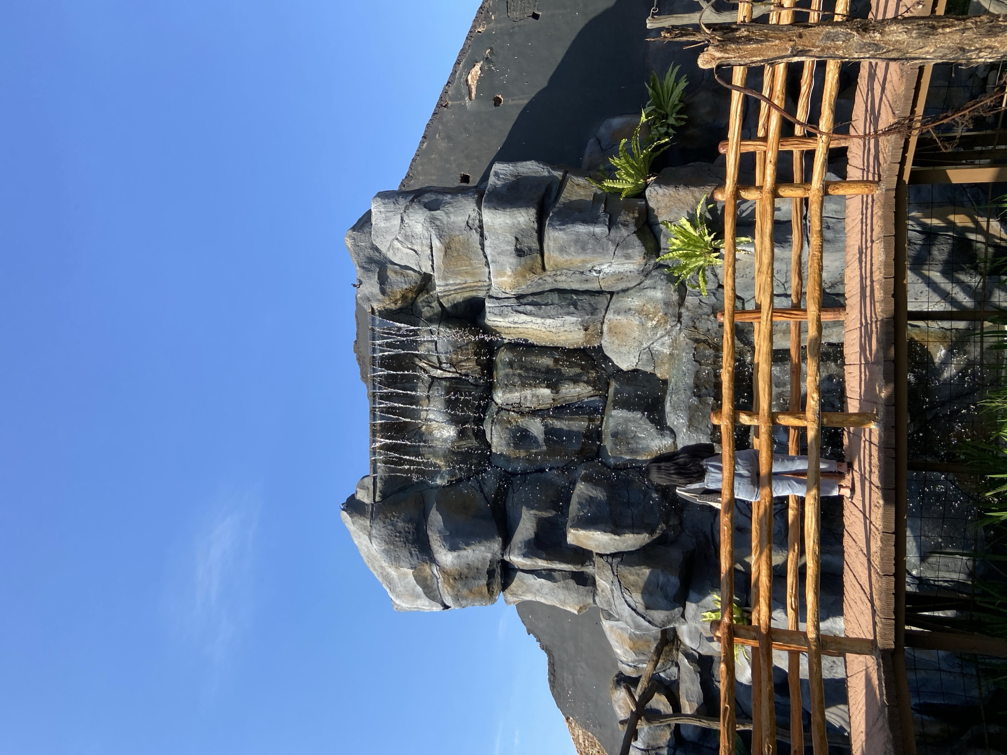 The scene is an outdoor environment in Mangli, Indonesia, depicting a rock waterfall feature next to a wooden bridge and fence. A person with long dark hair, wearing a light-colored top and denim pants, is standing on the bridge facing the waterfall. The artificial waterfall is constructed from stacked, grey-toned rocks and has plants growing out from some crevices. The sky is clear and blue with a few wispy clouds. The artificial rock formation is topped with a dark-colored, flat structure. The wooden bridge has a rustic fence made from upright logs.