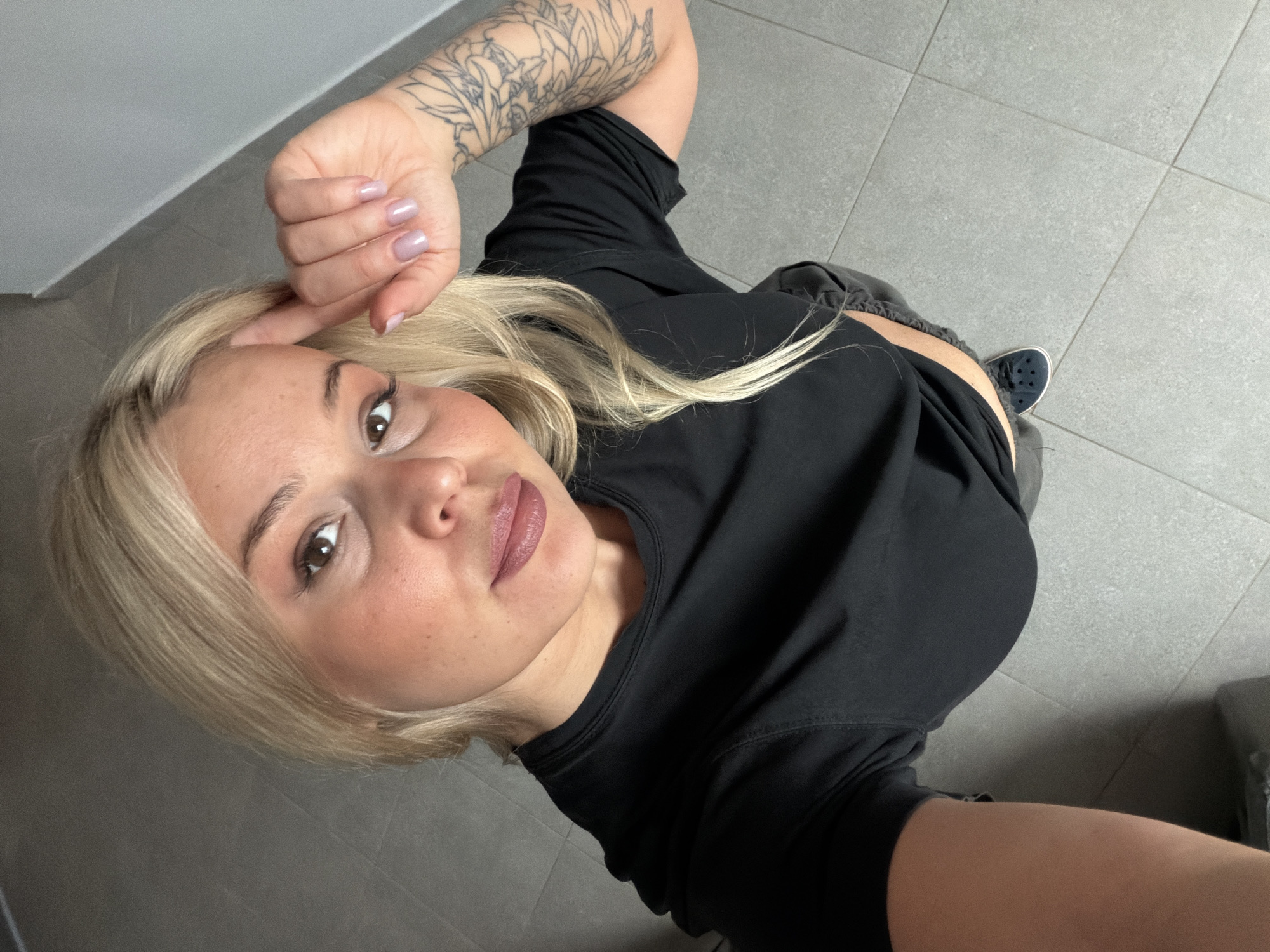 A fair-skinned woman with blonde hair and brown eyes is depicted in a high-angle selfie, looking directly at the camera with a subtle smile. She is wearing a black short-sleeved top and dark bottoms, possibly shorts, with dark blue slip-on shoes (resembling Crocs) visible on her left foot. Her right arm is bent, with her hand near her head, showcasing light purple nail polish on her manicured nails. A distinctive tattoo featuring a floral or leafy pattern covers a significant portion of her right forearm. The background consists of a light grey tiled floor and a plain light grey wall, suggesting an indoor setting. No specific time of day or weather conditions are discernible. The overall impression is casual and relaxed. No visible text is present in the image. The location context is Sarapul, Russia.