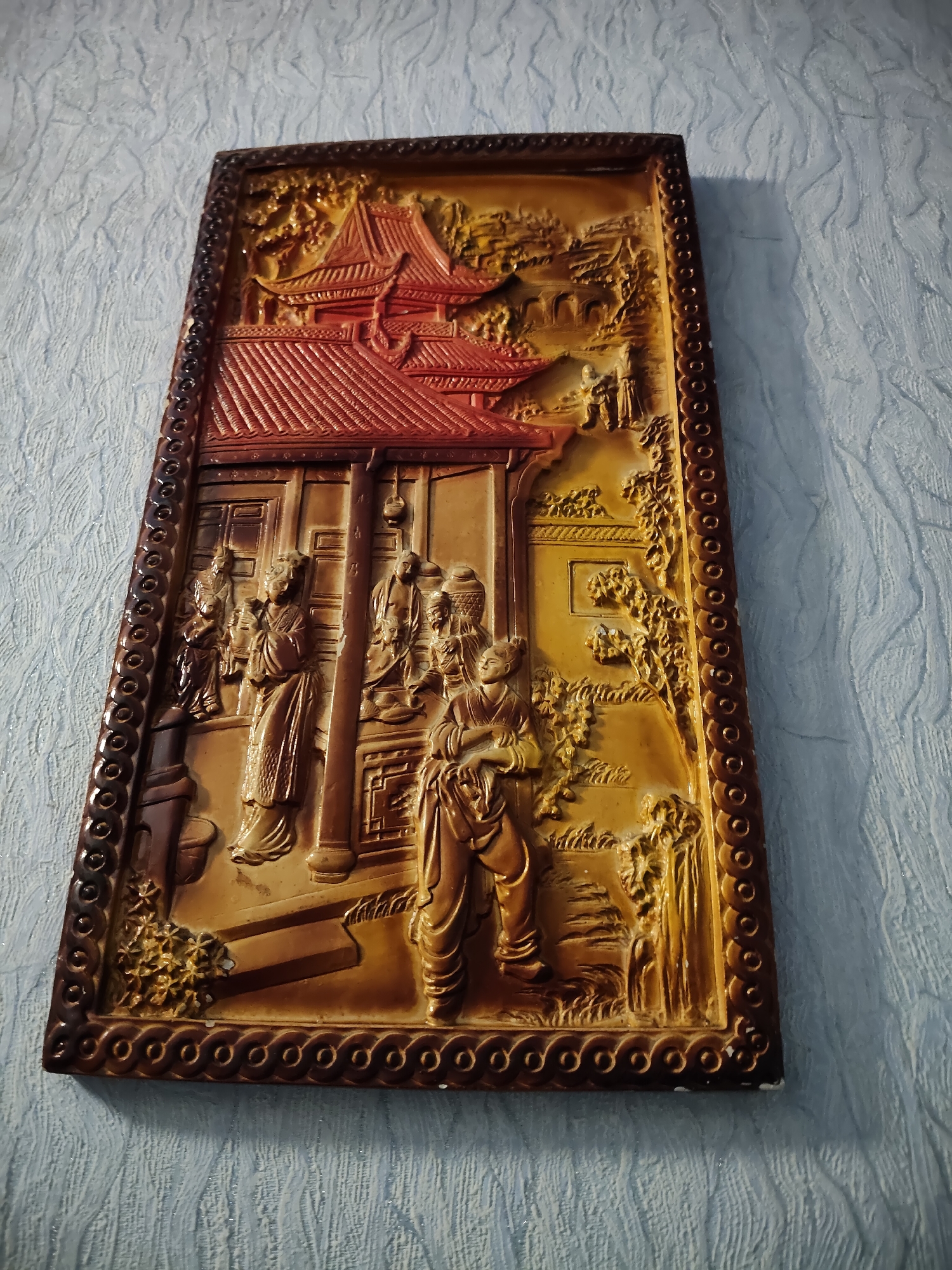 The image shows a rectangular, three-dimensional wall relief with an intricately carved scene depicting traditional East Asian architecture and figures. The relief is predominantly brown and gold, with a red tiled roof on the central building. The scene features several figures, possibly women in traditional garments, engaged in various activities within what appears to be a courtyard or garden setting. Lush foliage and a bridge are also visible. The relief is mounted on a textured, light blue wall. The specific location is not discernible from the image itself, and there is no information provided within the image or its description to indicate it is in Gîrbova, Moldova.