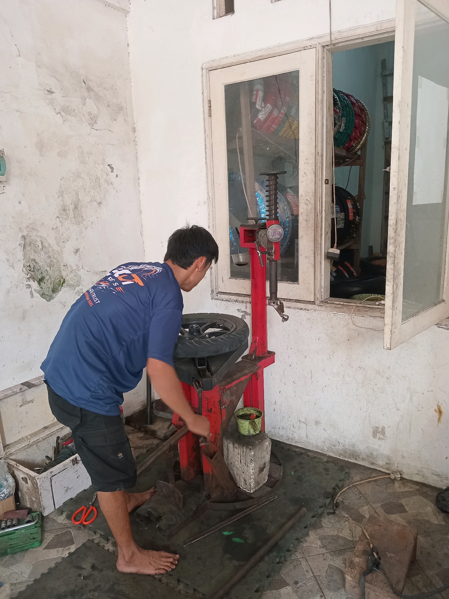 In Tebalo, Indonesia, a man is operating a red tire-changing machine inside a workshop. The man, wearing a blue t-shirt and black shorts, is barefoot and appears to be in the process of removing or installing a tire. The workshop environment shows a white wall with signs of wear and discoloration, and a window with multiple panes where tires are visible in the background. Tools and equipment, including a green toolbox and orange scissors, are scattered on the floor. The ground is a mix of patterned tiles and dark mats.