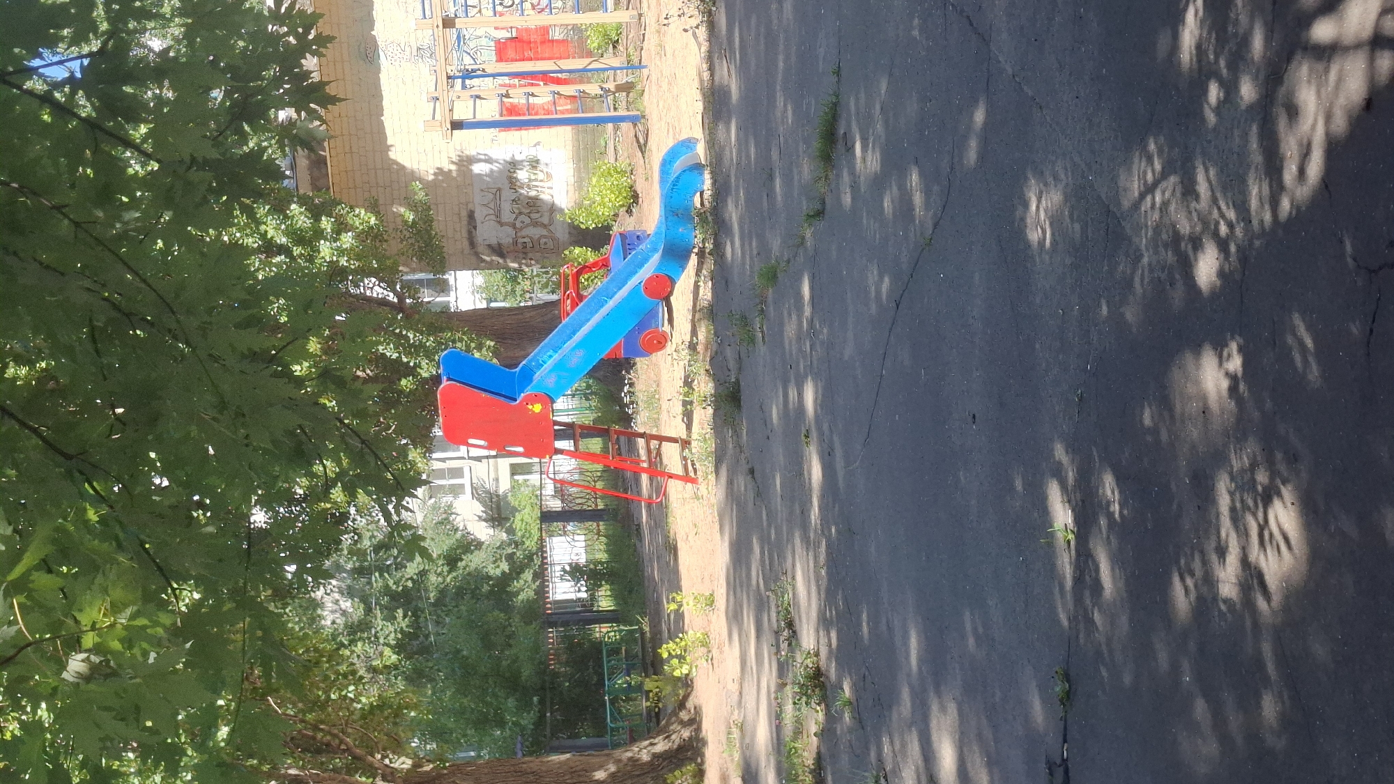 A red and blue plastic playground slide stands on a dirt and asphalt surface. The slide is elevated by a red metal ladder on the left side. The slide itself is blue, with a red top section that resembles a car's hood, complete with blue wheels. In the background, there is a brick building with graffiti on its wall, as well as trees and a metal fence. The ground is cracked asphalt with patches of dirt and sparse vegetation. Shadows from the trees are cast across the asphalt.