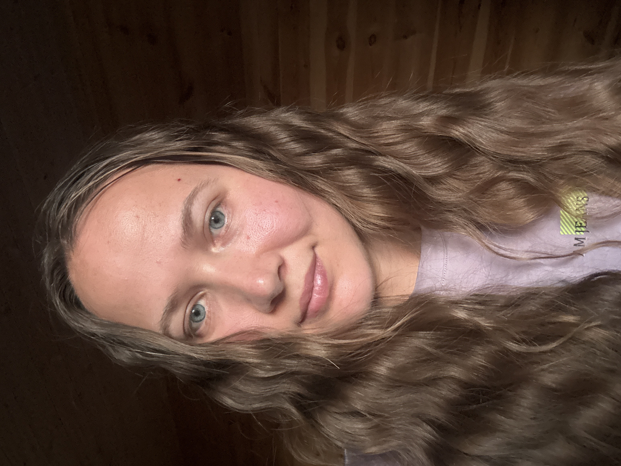 A close-up, rotated selfie of a young woman with long, wavy, light brown hair. She has fair skin, blue eyes, and a small red mark on her forehead. She is smiling slightly. She wears a light purple t-shirt with faded green text that reads "JEANS." The background appears to be a dark, possibly wooden, wall. No specific indicators suggest Tyulyuk, Russia.