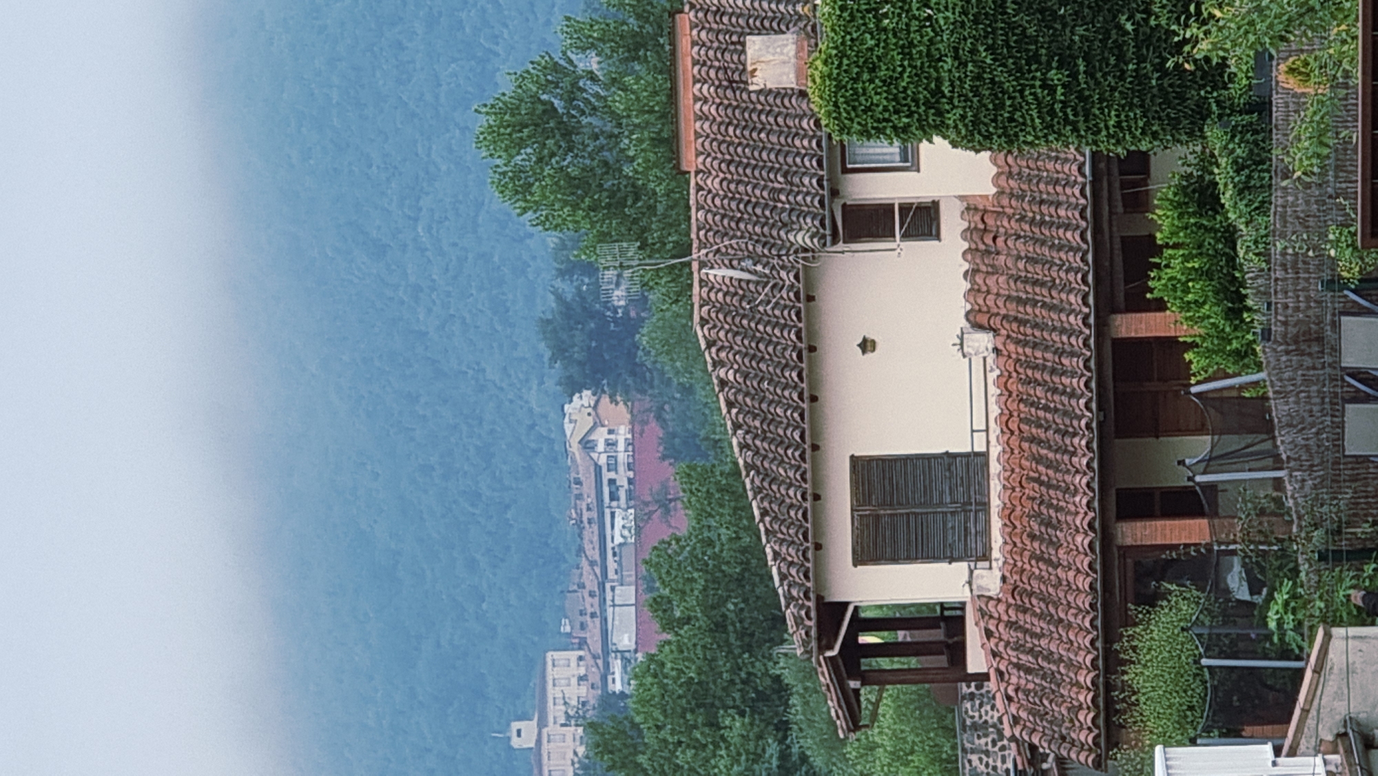 The image depicts a view of buildings and a forested mountainside in Sant Joan Les Fonts, Spain. In the foreground, a house with a terracotta tiled roof is partially visible. It has cream-colored walls, dark shutters on one window, and a chimney. Adjacent to this house, another section of a building with a similar tiled roof and brick pillars is visible. The middle ground shows a cluster of buildings with red tile roofs, some with white walls, and green trees. Further in the background, a larger building with a pale facade and a tower is situated on the slopes of a densely forested mountain. The mountain is covered in lush green trees and shrouded in a light mist or fog, creating a soft, hazy effect in the upper portion of the image. The overall scene conveys a sense of a residential area nestled within a natural, mountainous landscape.