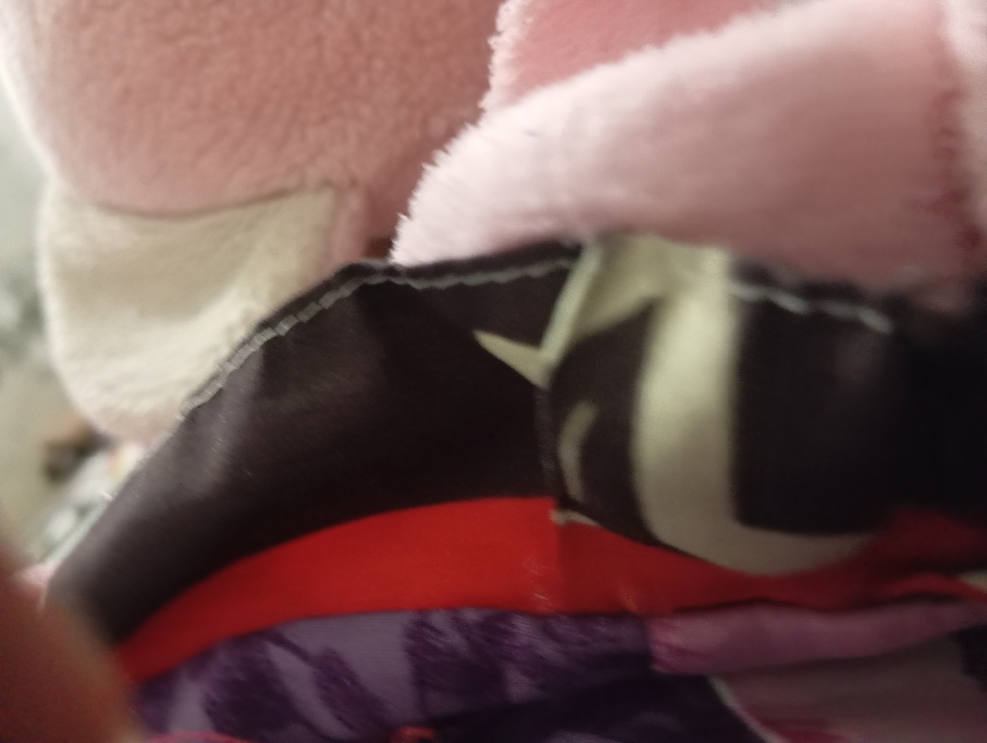 The image is a close-up, slightly blurry shot of fabric materials. In the foreground, a section of dark fabric with a white stripe pattern is visible. This fabric appears to be part of an article of clothing, possibly a jacket or backpack, given the stitching and layered appearance. To the left of this dark fabric, there are sections of brightly colored fabric, including a vibrant red and a purple fabric with a patterned design. To the right, a soft, pink fuzzy material is prominent, also suggesting it's part of a textile object. The background is out of focus, but hints at other soft textures and potentially a blurred human hand or arm in the upper left corner. The scene has a domestic or personal feel due to the nature of the materials. There is no indication of the city of Rejosari, Indonesia, within the image itself.