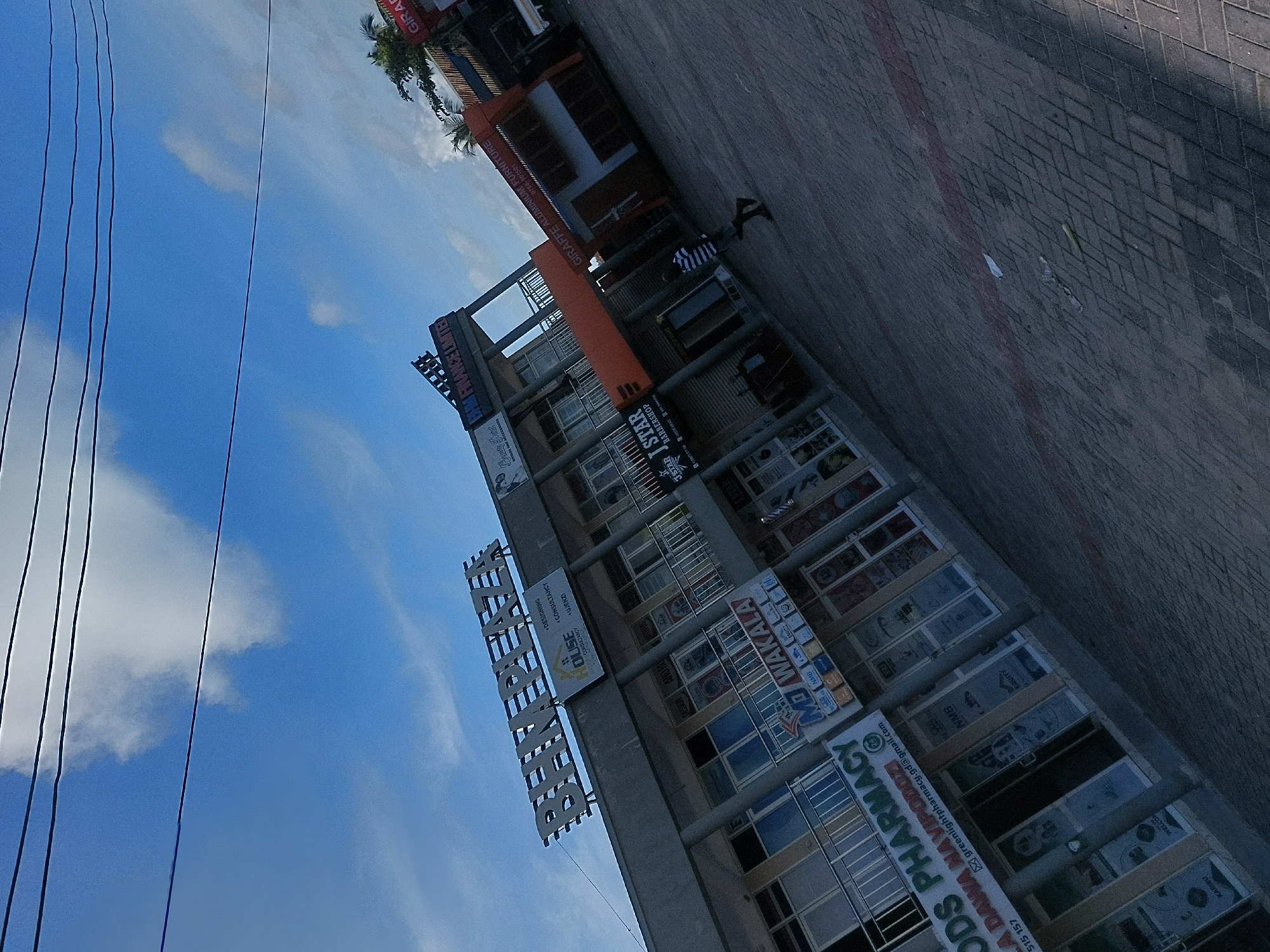 The image shows a multistory commercial building in Dar es-Salaam, Tanzania, named "Binetha Plaza." The building houses various businesses, including the "Ods Pharmacy," "MP Wakala," "Star I Star Barbershop," and "Giraffe Aluminium Furniture." The building has a sign for "Bali Finance Limited." A person is visible walking in front of the building on a paved walkway. Power lines are in the sky. The sky is blue with some clouds.