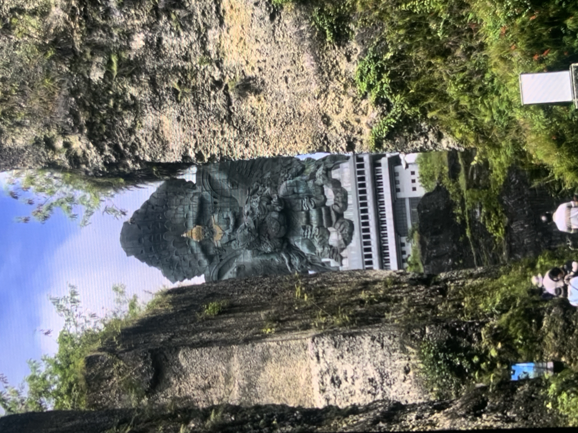 The image shows a large stone statue of a deity, possibly Hindu, with multiple arms and a headdress, positioned between two steep, rocky cliffs covered in vegetation. The statue is made of a dark, greenish material and is quite detailed. Below the statue, a building with a patterned facade, resembling a row of windows or architectural details, is visible. The environment appears to be a natural or man-made canyon, with lush green plants and trees growing on the rocky slopes. In the foreground, there are a few people walking and standing, some of whom are near a sign or display. The sky is visible above the statue, and it appears to be a bright day. This image does not depict Stavropol, Russia, but rather a location that appears to be a religious or cultural monument set within a dramatic natural landscape.