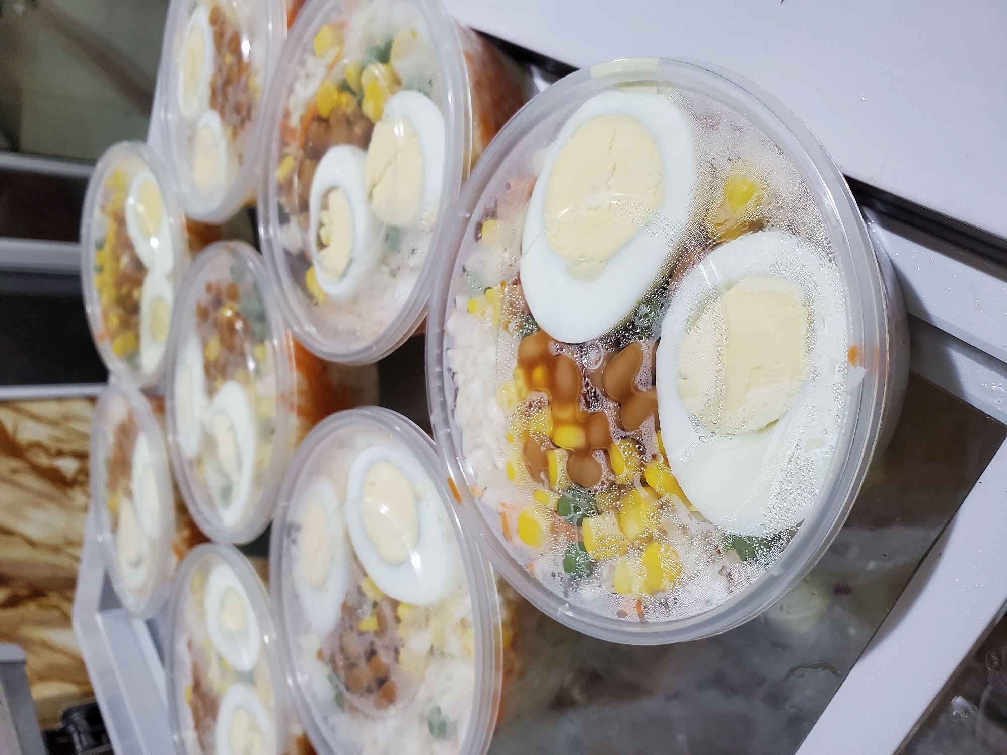 The image shows a collection of seven clear plastic containers filled with food. Each container appears to hold a similar meal, featuring rice, vegetables like corn and peas, small brown nuggets (possibly fried or roasted), and sliced hard-boiled eggs on top. The containers are arranged close together on a reflective white surface, which could be a countertop or a tray. The lids of the containers are slightly fogged with condensation. The image was taken in a location consistent with Nkpor, Nigeria.