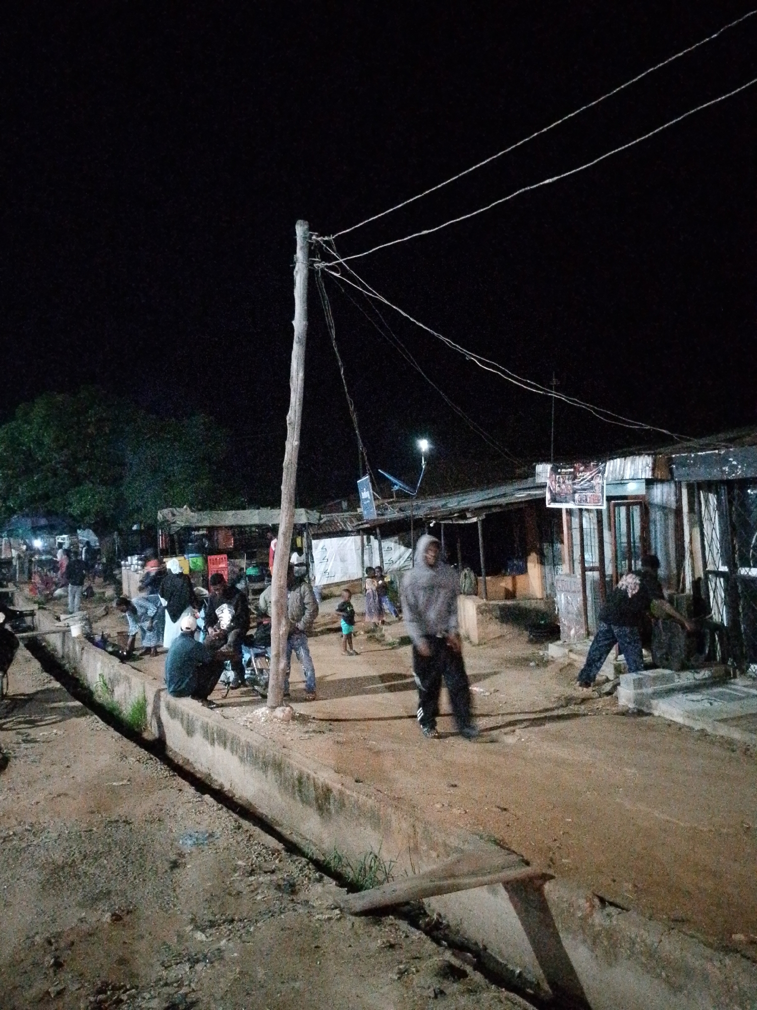 In the city of Unknown, Unknown, at night, a group of people are gathered in an outdoor area that appears to be a market or informal settlement. A wooden pole stands in the foreground, supporting electrical wires that stretch across the frame. Several corrugated metal structures are visible in the background, suggesting makeshift shops or dwellings. People are seen standing, sitting, and interacting. One person in a grey hoodie is in the foreground, facing left. Another person in a black shirt is in the mid-ground, bent over an object. A concrete drainage ditch runs along the left side of the scene, filled with dirt and some vegetation. The ground is predominantly dirt and gravel. The lighting is dim, likely from artificial sources not fully visible.