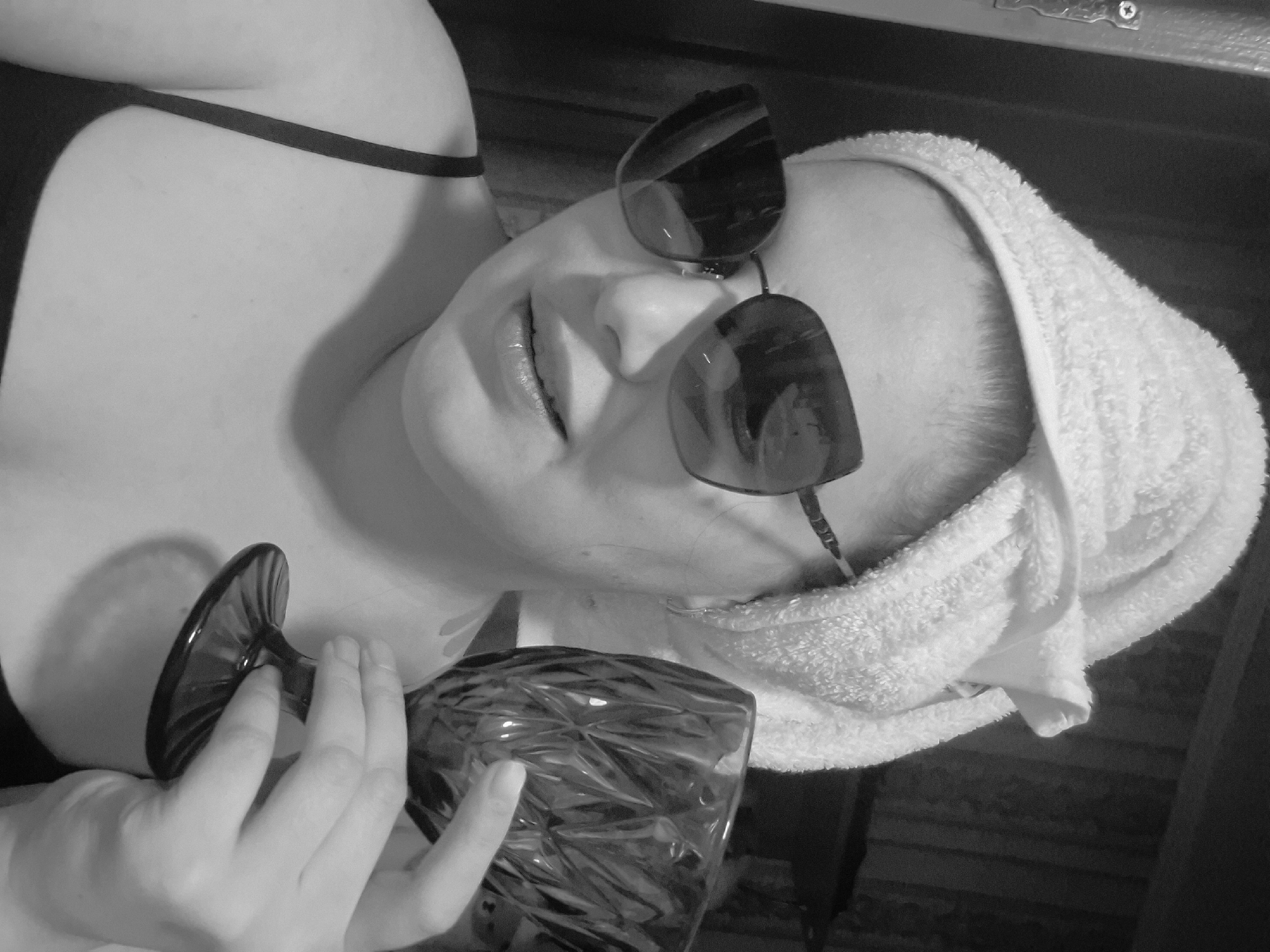 A black and white photograph, captured in Chaikovsky, Russia, depicts a person reclining, viewed from a high angle. The subject, likely female, is lying down with their head wrapped in a white, fluffy towel. Dark sunglasses rest on their face, covering their eyes, and their lips are slightly parted. A dark bra strap is visible over their shoulder. In the foreground, the person's hand holds a dark, textured glass, possibly a goblet or large wine glass. The background is indistinct and dark, suggesting an indoor or dimly lit environment. The image conveys a sense of relaxation or a posed moment. No specific time of day, weather, or visible text can be determined from the image.
