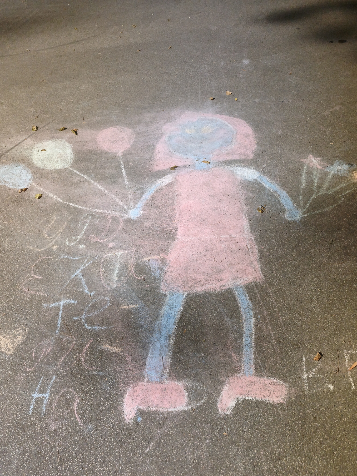 The image shows a chalk drawing on asphalt, likely in a public area such as a park or sidewalk, in the city of Lovech, Bulgaria. The drawing depicts a childlike figure with a pink dress and blue limbs and head, holding three balloons. One balloon is white, another is pink, and the third is blue, all rendered as circles with lines extending from them. The figure's arms are raised, with one holding the pink and blue balloons and the other extended to the side, holding a fan-like object with lines radiating outward.
Beneath the figure, there is chalk writing in a script that appears to be Cyrillic. Parts of the writing are difficult to decipher due to the chalk's uneven application and the texture of the asphalt, but visible characters and segments suggest words or names. Some visible letters include "У", "Д", "Е", "К", "Т", "Е", "Н", "Н", "О", "Н", "Н", "А", and "Е". The drawing is in pastel chalk colors: pink, blue, and white. Fallen leaves are scattered on the asphalt around the drawing, suggesting an outdoor environment and possibly an autumn season. The lighting suggests it is daytime, with some shadows visible, possibly from trees or structures.
