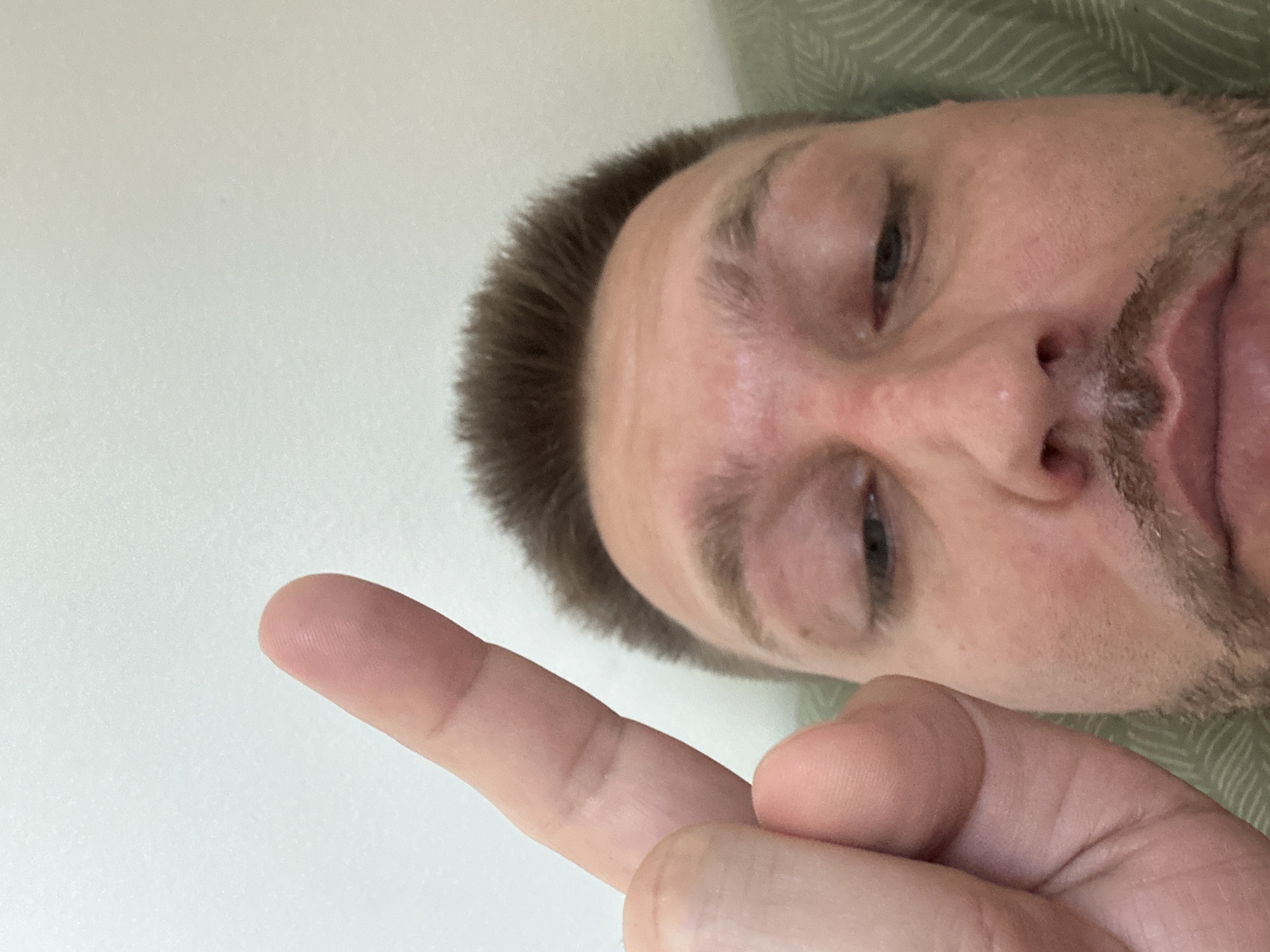 A close-up, low-angle selfie shows a man with short brown hair and light-colored stubble. His right index finger is raised towards the camera, obscuring part of his forehead and eye. His eyes are looking down, and his mouth is slightly open. The background is a plain, light-colored wall and a portion of a green patterned surface, possibly a pillow or blanket. There is no discernible information about the location in the city of Zvezdara Urban Municipality, Serbia, within the image.