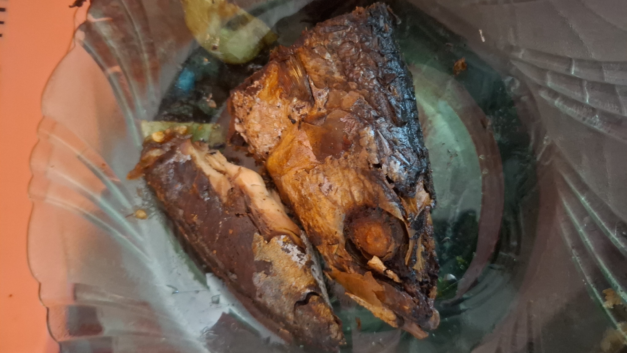 The image shows two pieces of fried fish in a clear, fluted glass bowl. The fish are dark brown and appear to be somewhat dry, with some visible scales. One piece is larger and positioned in the foreground, while the other is smaller and behind it. There are hints of green vegetables, possibly peppers or limes, peeking out from under the fish. The bowl is resting on an orange surface, which is slightly out of focus. The lighting is overhead, casting shadows within the bowl. The scene appears to be a close-up of a meal, likely prepared at home.