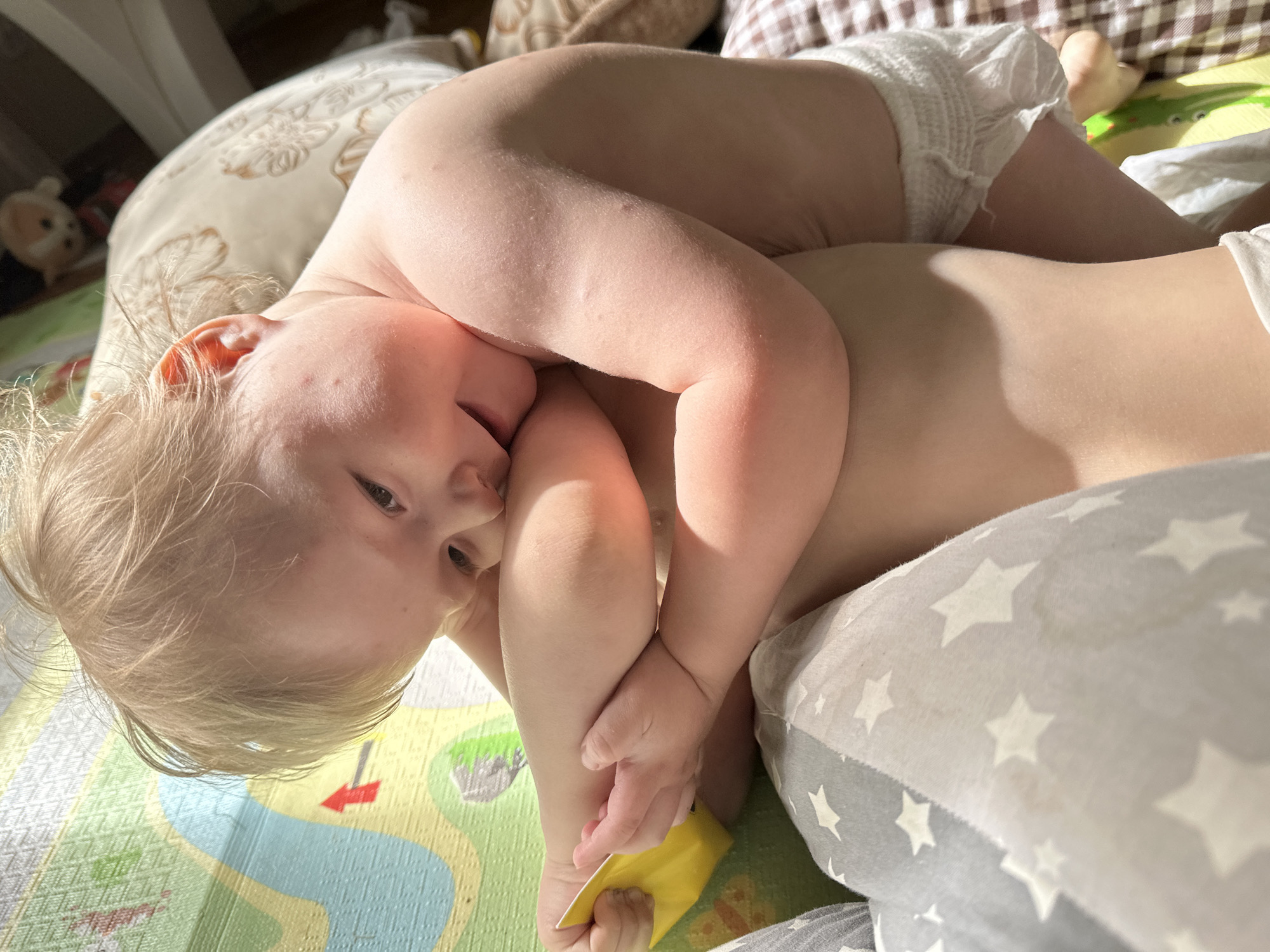 A close-up shot captures two young children lying on what appears to be a playmat with a cartoon river and other colorful designs. One child is wearing a diaper and lying on their back, while the other child, also wearing a diaper, is lying on top of them. The upper child's arm is wrapped around the lower child. The room is filled with natural light, and pillows are seen in the background. A corner of a blanket with star patterns is visible in the bottom right corner. The photograph does not show any signs indicating the location in the city of Nizhny Novgorod, Russia.