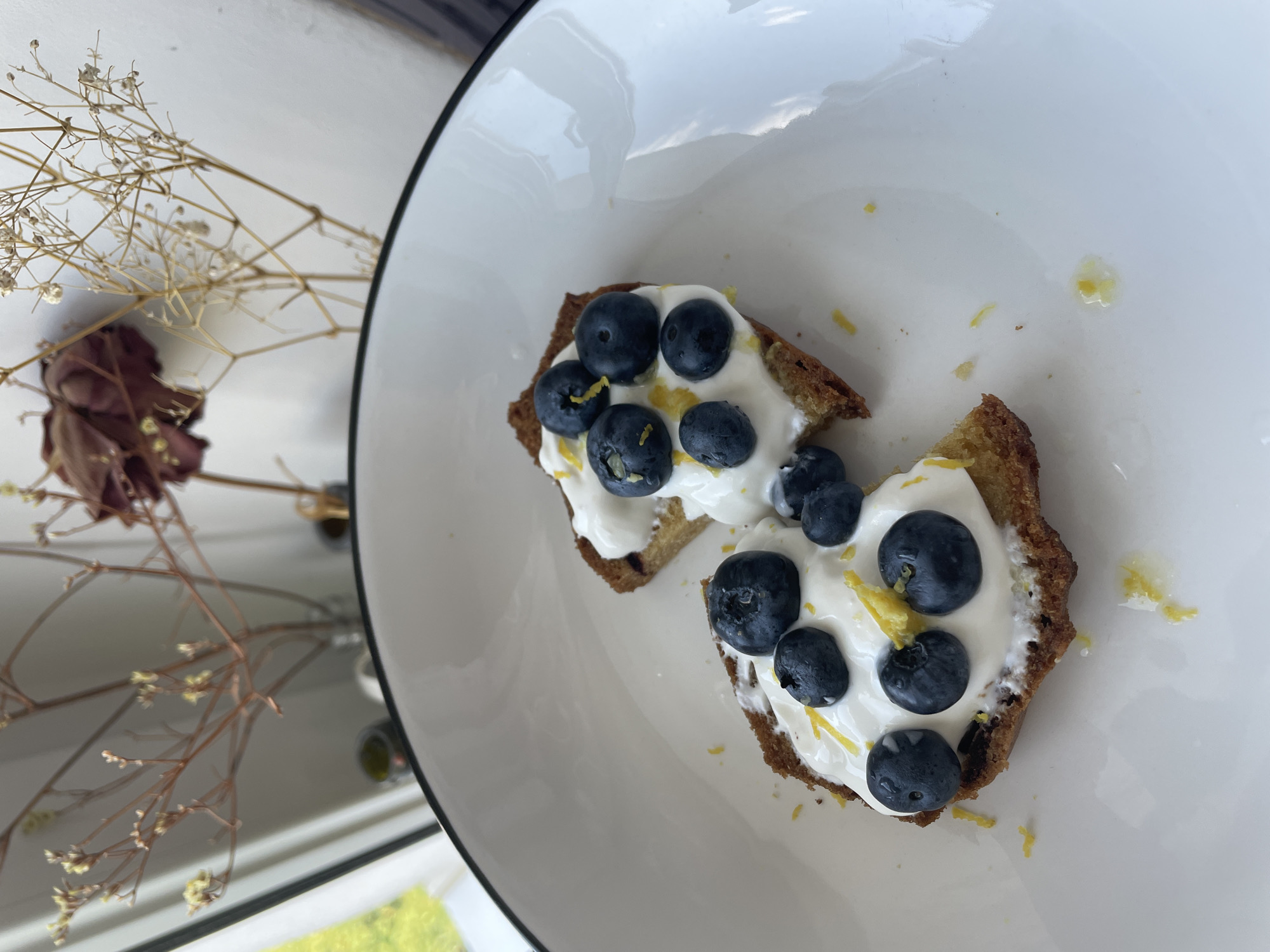 The image shows two pieces of toast on a white plate. Each piece of toast is topped with a white spread, blueberries, and what appears to be lemon zest. The toast is golden brown and appears to be somewhat crumbly. In the background, there are dried flowers in vases, and a window with greenery visible outside. The overall scene suggests a breakfast or snack setting. There is no information in the image to indicate a specific location in the city of Hrodna, Belarus.