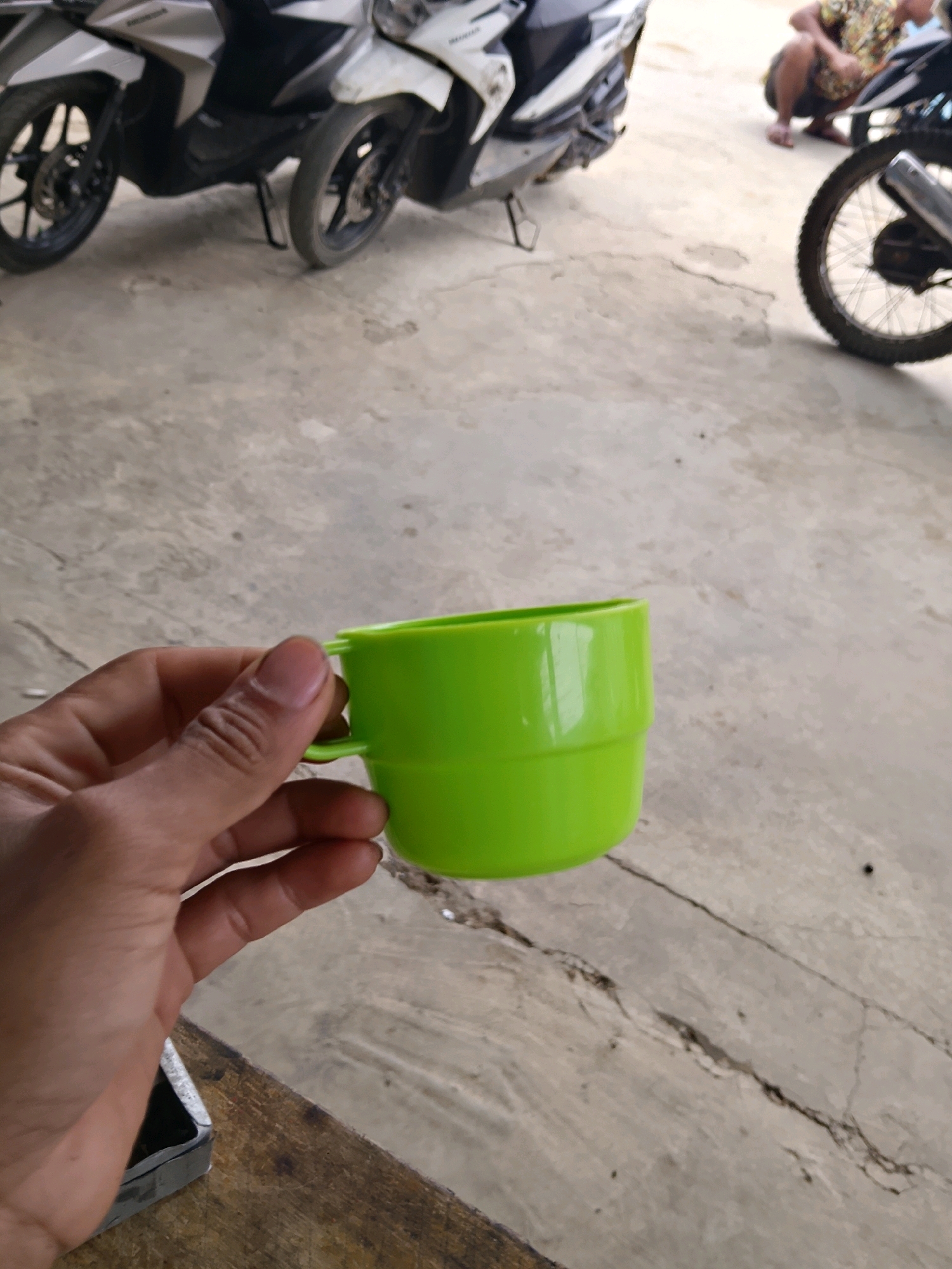 A person's hand is holding a bright green plastic cup on a concrete surface. In the background, two white and black scooters are parked, and a person is crouched down next to one of them. The scene appears to be outdoors, possibly in a parking lot or a street in an urban environment. The lighting suggests it is daytime.