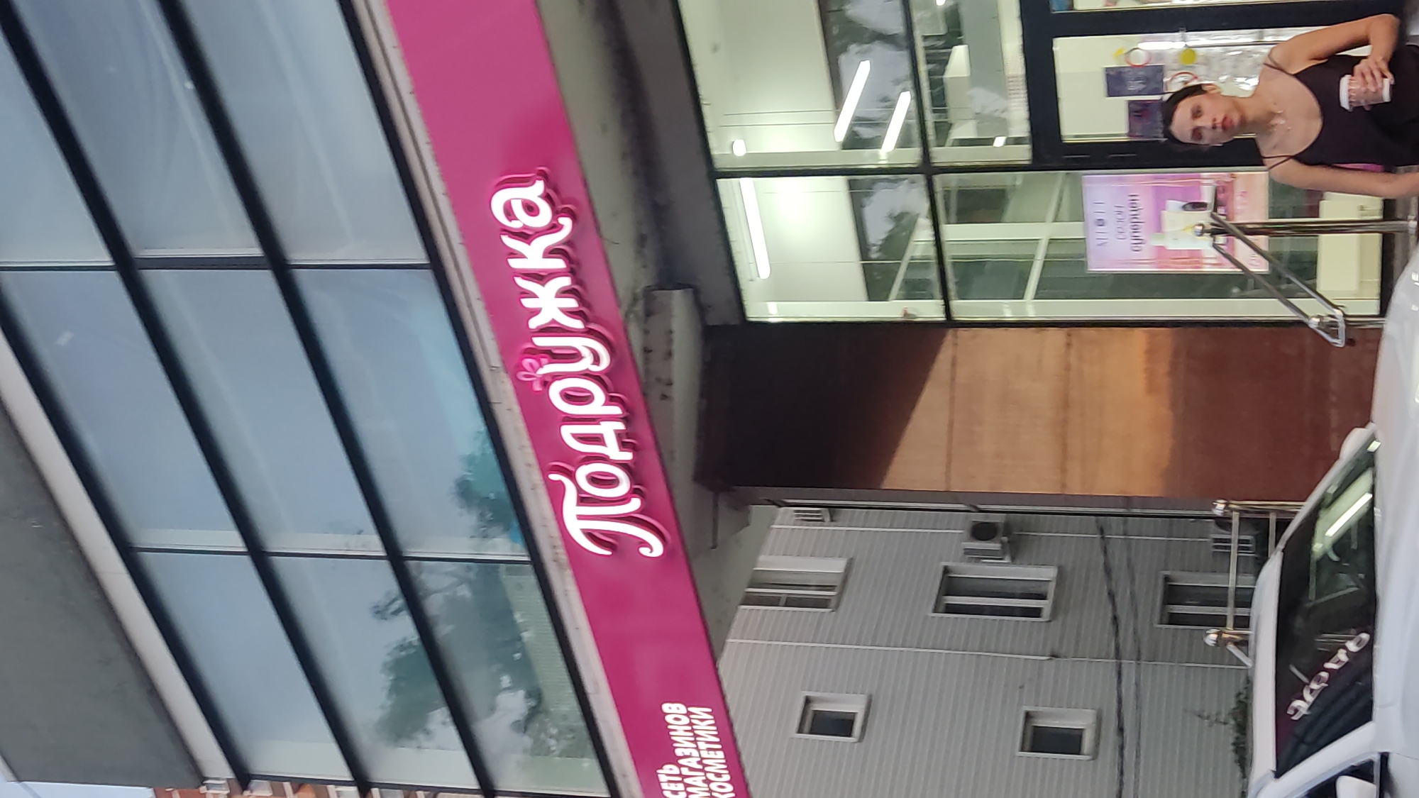 The image shows a street-level view of a building with a bright pink sign in Russian. The sign reads "Подружка" which translates to "Girlfriend" or "Little friend". Below this, there is smaller text that is partially obscured but appears to read "СЕТЬ МАГАЗИНОВ КОСМЕТИКИ", meaning "Chain of cosmetic stores." The building has large windows on the upper floors, reflecting the sky and some trees. A white car is parked in the foreground, and a woman in a dark tank top is walking in front of the store entrance, holding a coffee cup. The building's facade is modern with clean lines. The overall setting suggests a commercial area in a city. Krasnodar, Russia is not explicitly mentioned in the image, but the Russian signage indicates it is likely in Russia.