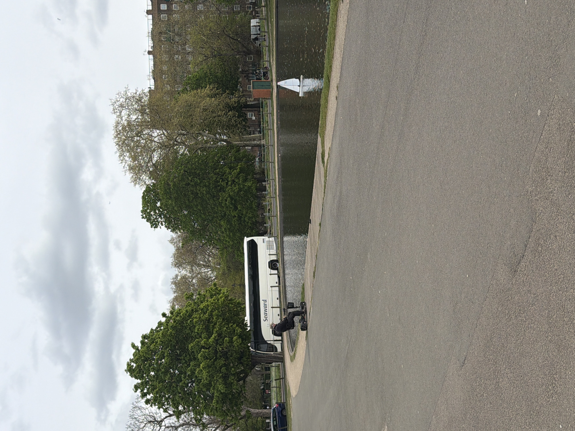 A horizontal image shows a paved path with a canal to the left. A large coach bus is parked alongside the canal bank. A person sits on a mobility scooter beside the bus, near the canal edge. A model sailboat floats in the canal. In the background are trees and a multistory building. The sky is overcast. This scene is likely in one of London's Royal Parks.