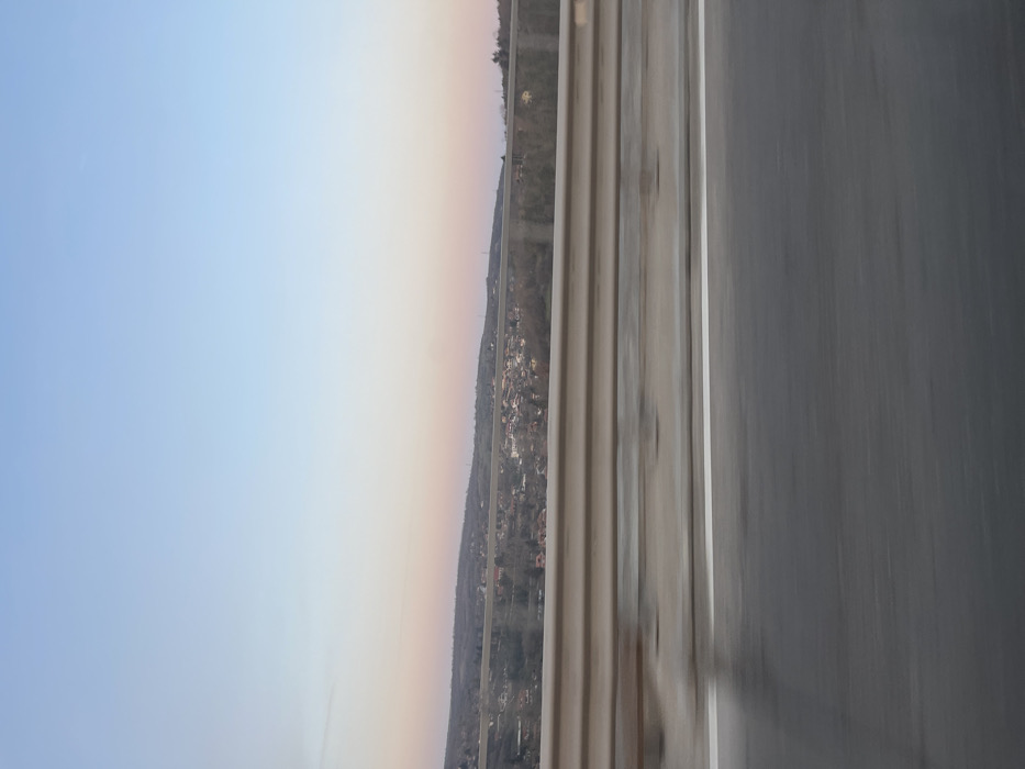 The image displays an elevated view of the settlement of Pisanets, Bulgaria, observed from a moving vehicle. In the immediate foreground, a blurred dark asphalt road surface is present, with a distinct white solid line visible on the right. Above the road, a two-tier metal guardrail, light gray in color and exhibiting motion blur, spans horizontally across the frame. Beyond the guardrail, a valley contains a scattered collection of residential structures featuring reddish-brown roofs, interspersed among a landscape dominated by bare deciduous trees and some evergreen foliage. This settlement extends across undulating and hilly terrain. The background consists of rolling hills covered in sparse, dark vegetation. The sky overhead transitions from light blue to a pale pink or peach hue near the horizon, suggesting conditions consistent with dawn or dusk. No individuals are visible.