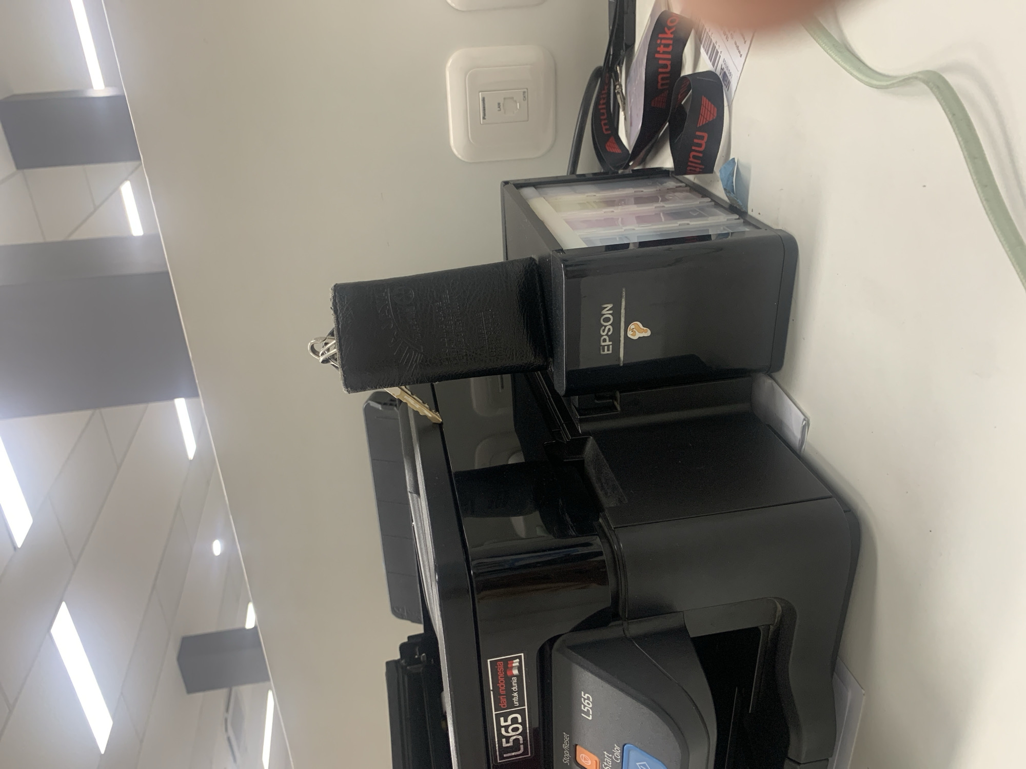 The image depicts a close-up view of a black Epson L565 printer situated on a white desk, likely in an office environment. A black leather keychain with a metallic ring is placed on top of the printer's ink cartridge housing. The printer has visible labels, including "L565" and "dari indonesia untuk dunia" with small Indonesian flags. To the right of the printer, a white wall-mounted LAN port is visible, along with a black lanyard with "multitika" branding. The scene is illuminated by overhead fluorescent lights, suggesting an indoor setting. The image does not provide specific details that would allow for the determination of its exact location within the city of Palembang, Indonesia.