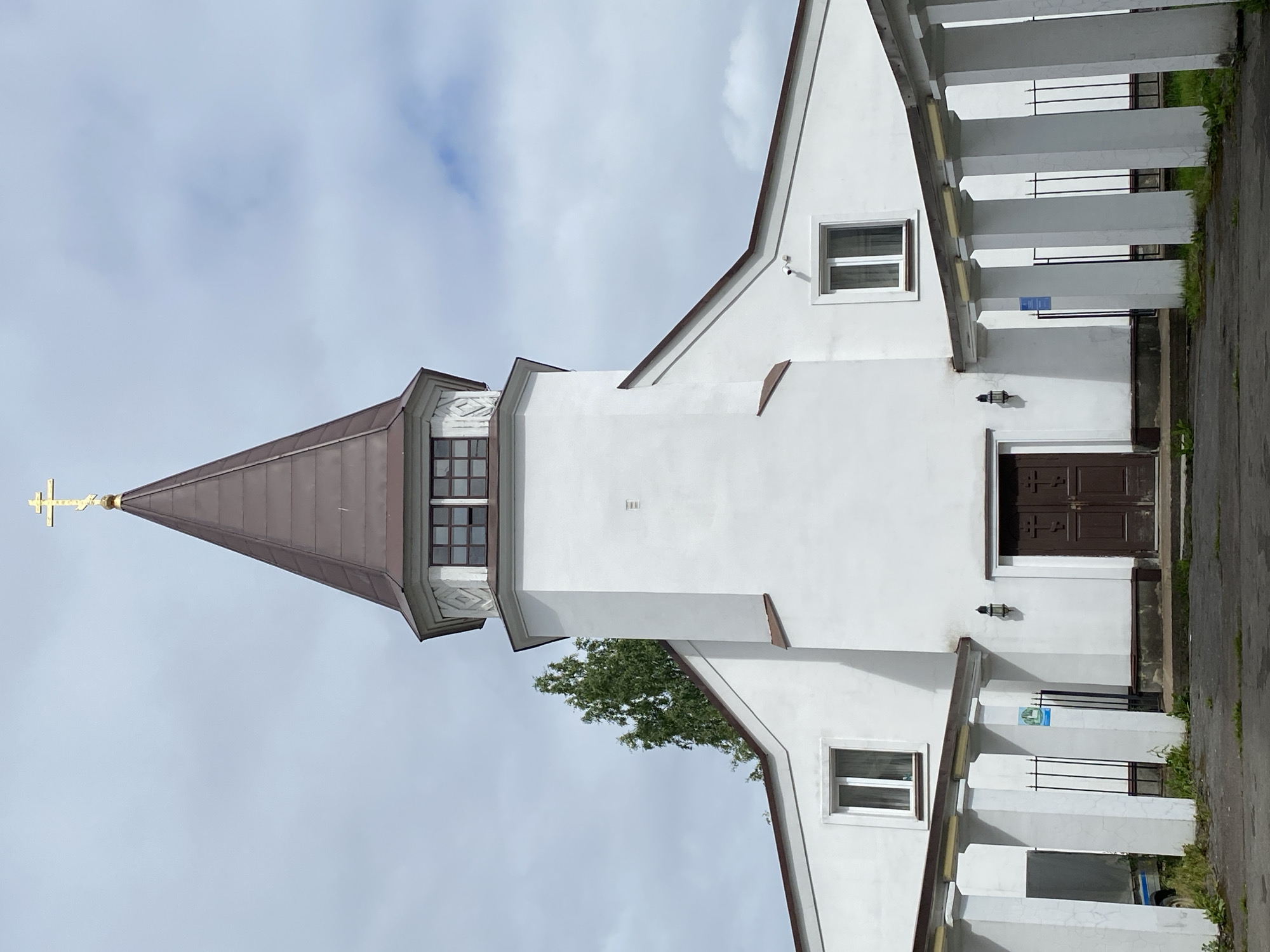 The image shows the exterior of a white church with a brown, conical steeple topped by a gold cross. The church has a prominent entrance with double brown doors. There are two windows on either side of the main entrance, under the eaves of the building. A white colonnade with a slanted roof runs along the front of the building. The sky is cloudy and overcast. The church is located in Sortavala, Russia.