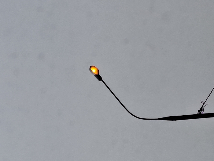 A single street light, located in Obukhovo, Russia, is depicted against an overcast, light grey sky. The dark, slender pole of the street light extends upwards from the lower left portion of the frame, supporting a dark, curved arm that culminates in an elongated, tear-drop shaped lamp housing. The lamp is actively illuminated, emitting an orange-yellow light. Near the base of the pole, several dark wires, some exhibiting faint purple hues, are visible and appear to be secured or tangled. No other structures, individuals, or environmental elements are discernible within the frame.