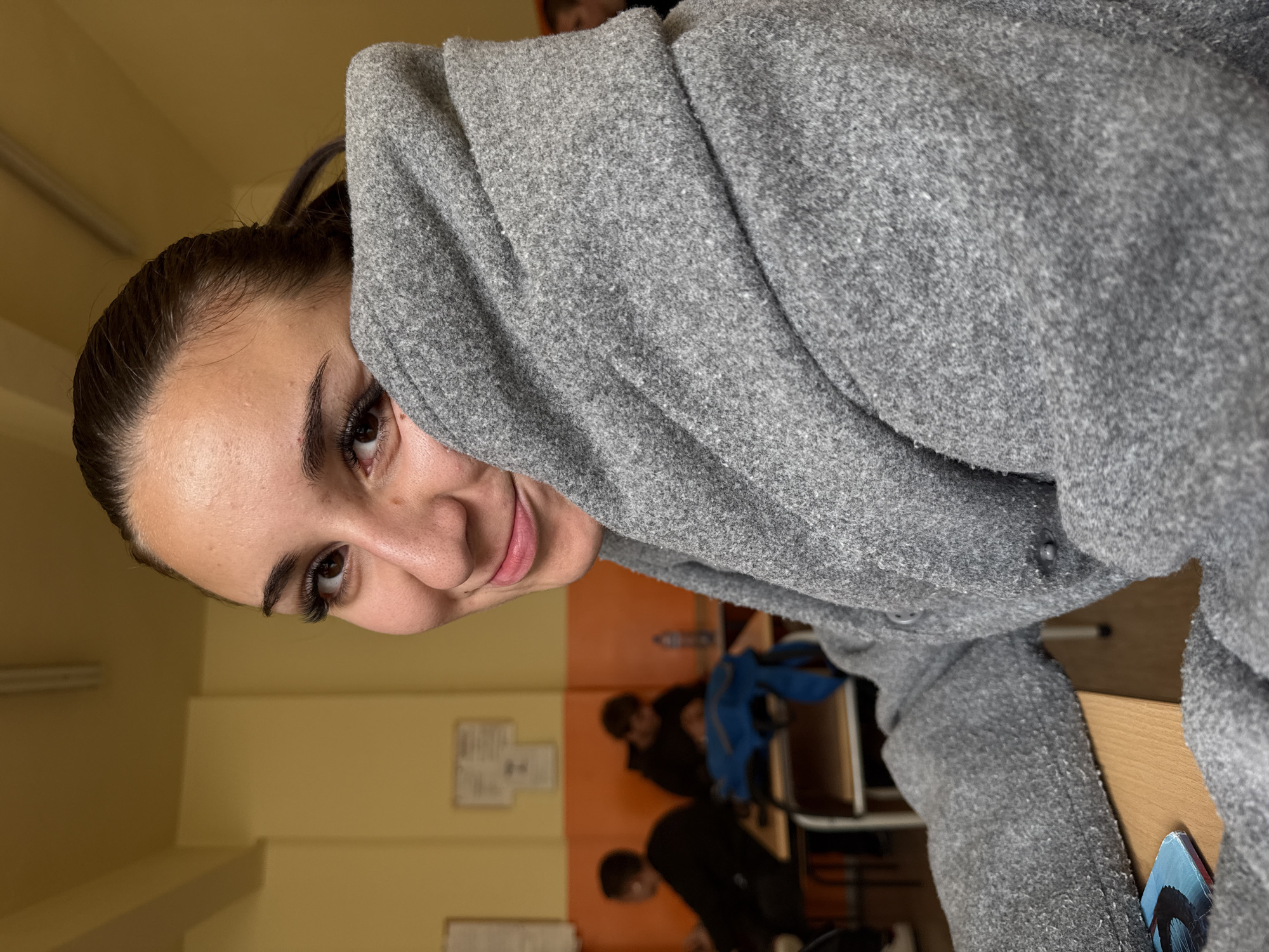 The image shows a young woman with dark hair pulled back, wearing a grey coat with the hood pulled up, partially obscuring her face. She is smiling and looking towards the camera. The background indicates an interior space, possibly a classroom, with other people seated at desks. The walls are painted yellow and orange. There are papers hanging on the wall. Bottles are placed on a surface in the background. The location in the city of Sofia, Bulgaria, cannot be determined from the image.