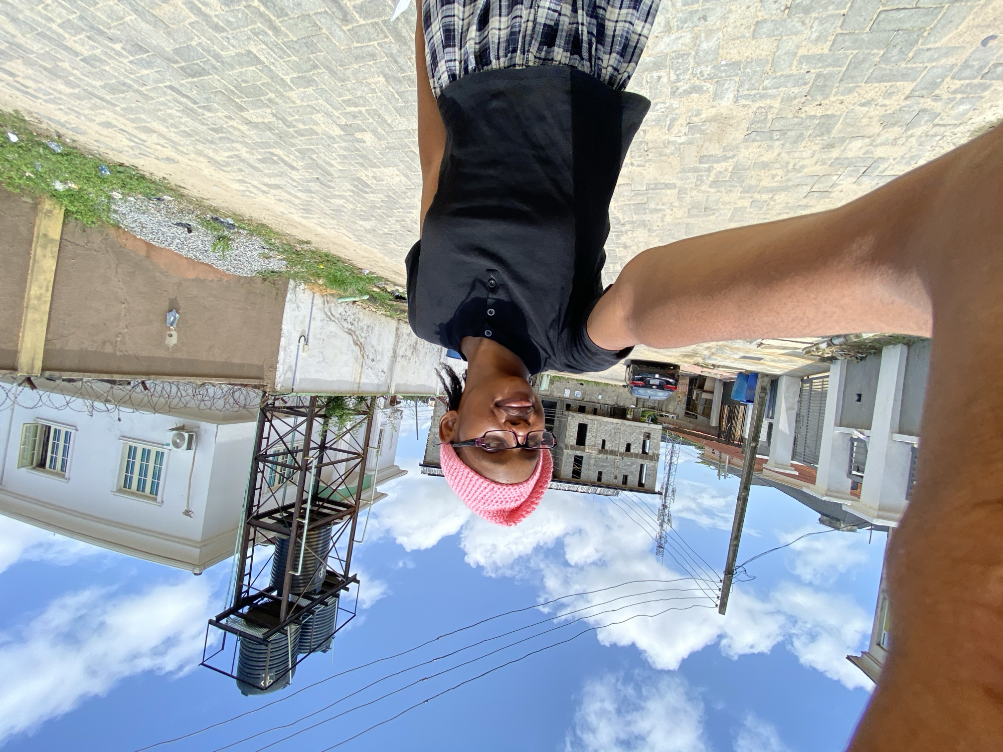 A person wearing glasses and a pink hat is visible in the foreground, taking a selfie. They are wearing a black top and a plaid skirt. The background shows a street with buildings, including a white building with a water tank and a building under construction. Utility poles and wires are also visible against a blue sky with clouds. The image appears to be taken in Unknown, Unknown.