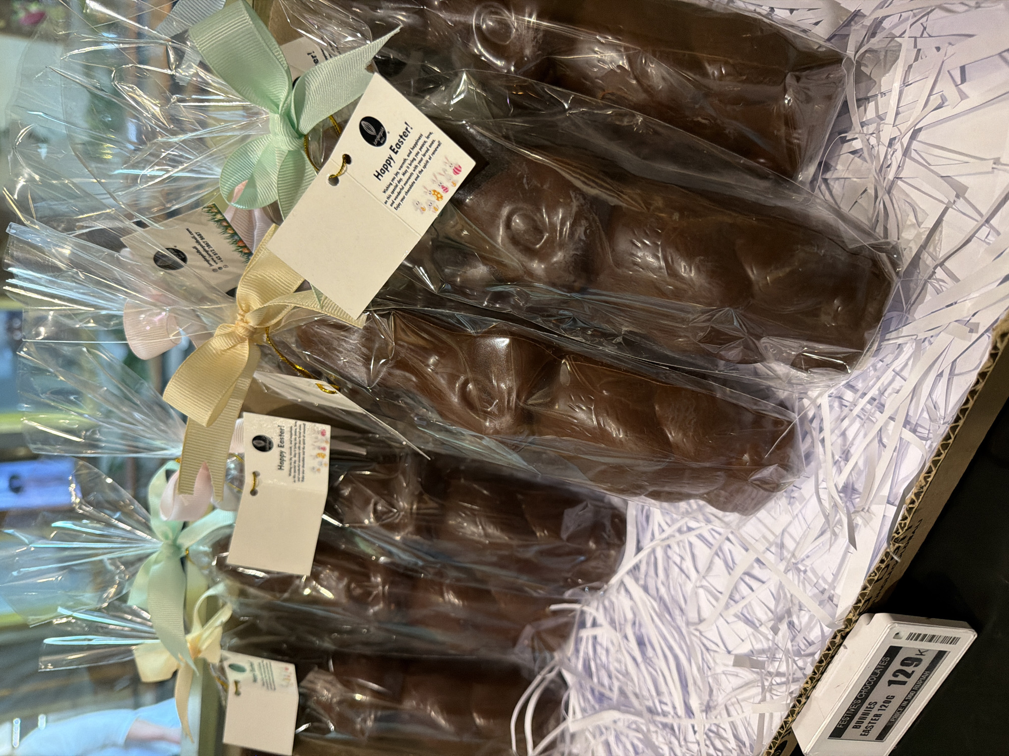 The image shows a box of Festive's Chocolates Easter Bunnies, priced at 129K (likely Indonesian Rupiah), weighing 120g. The chocolates are individually wrapped in clear plastic and tied with ribbons and "Happy Easter!" tags from Jungle Gold. The chocolates are dark in color and shaped like bunnies. The box is lined with white shredded paper. Based on the information on the tags, these chocolates are likely available in Mekarsari, Indonesia.